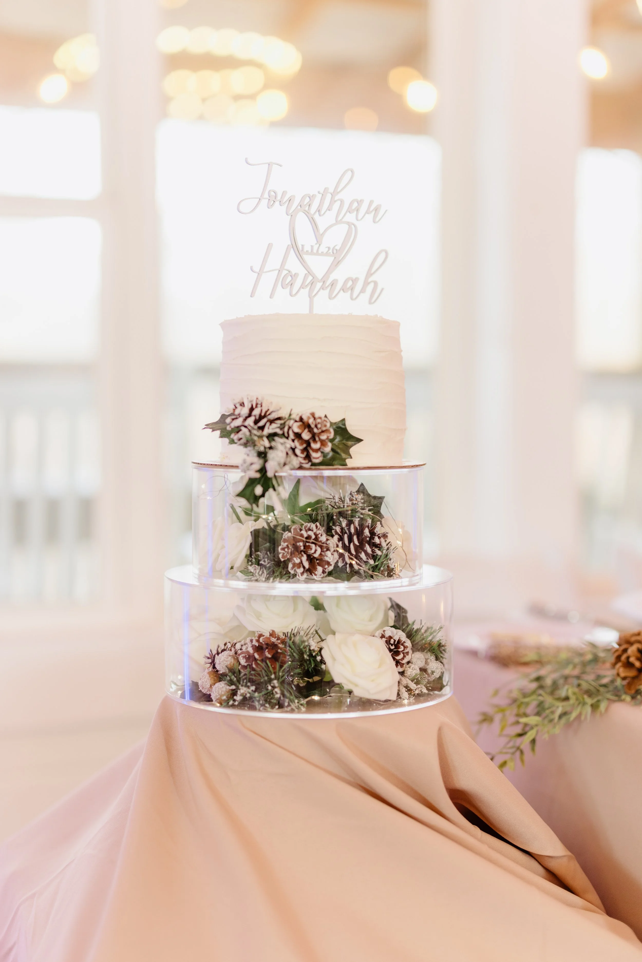 A wedding cake with a white textured frosting, decorated with pinecones and white roses, topped with a silver cake topper reading 'Jonathan & Hannah' with a heart and date, placed on a three-tier transparent display with pink fabric draped underneath