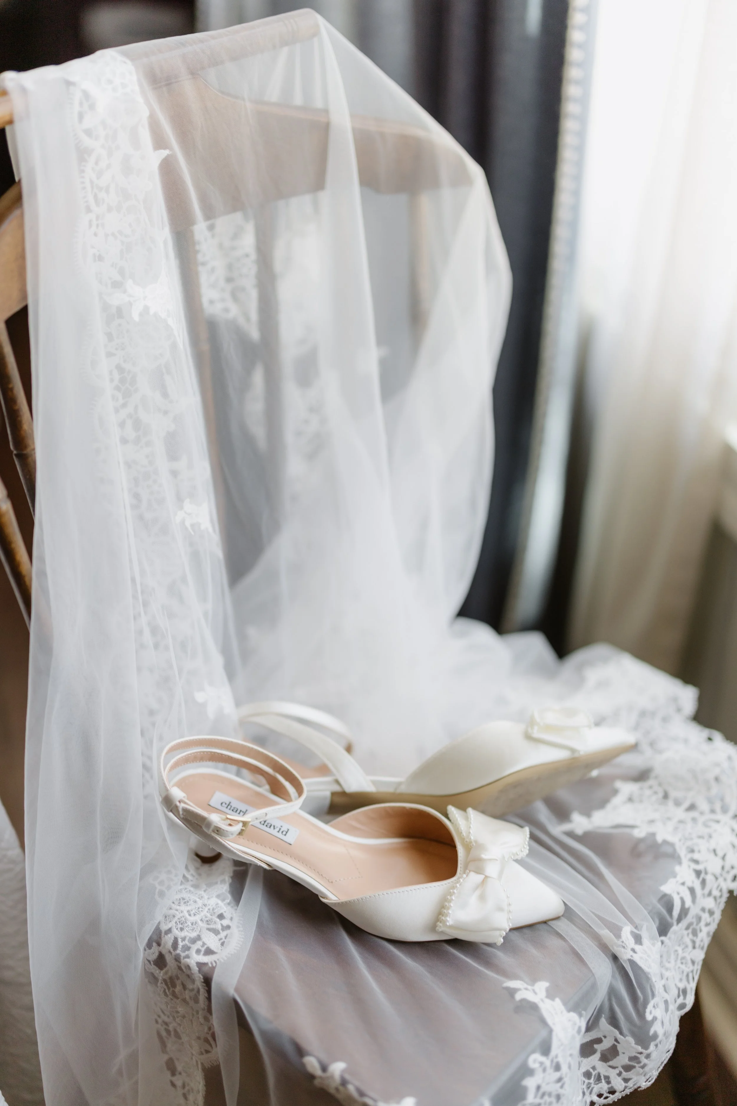 White wedding shoes with bows and a veil with lace detail on a lace-draped chair near a window.