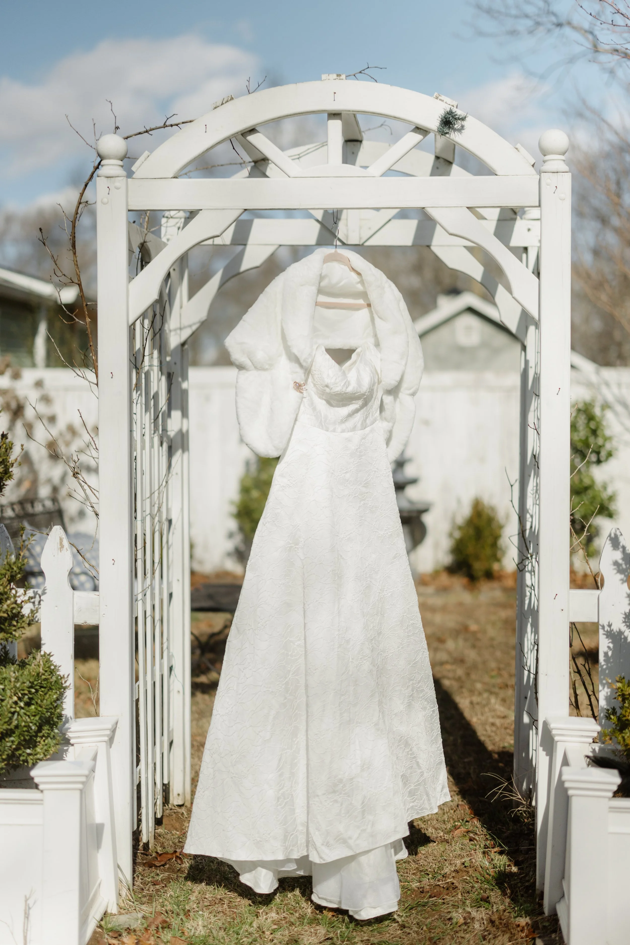A white wedding dress with a matching white furry shawl hanging on a hanger, displayed on a white arch outdoors on a sunny day.