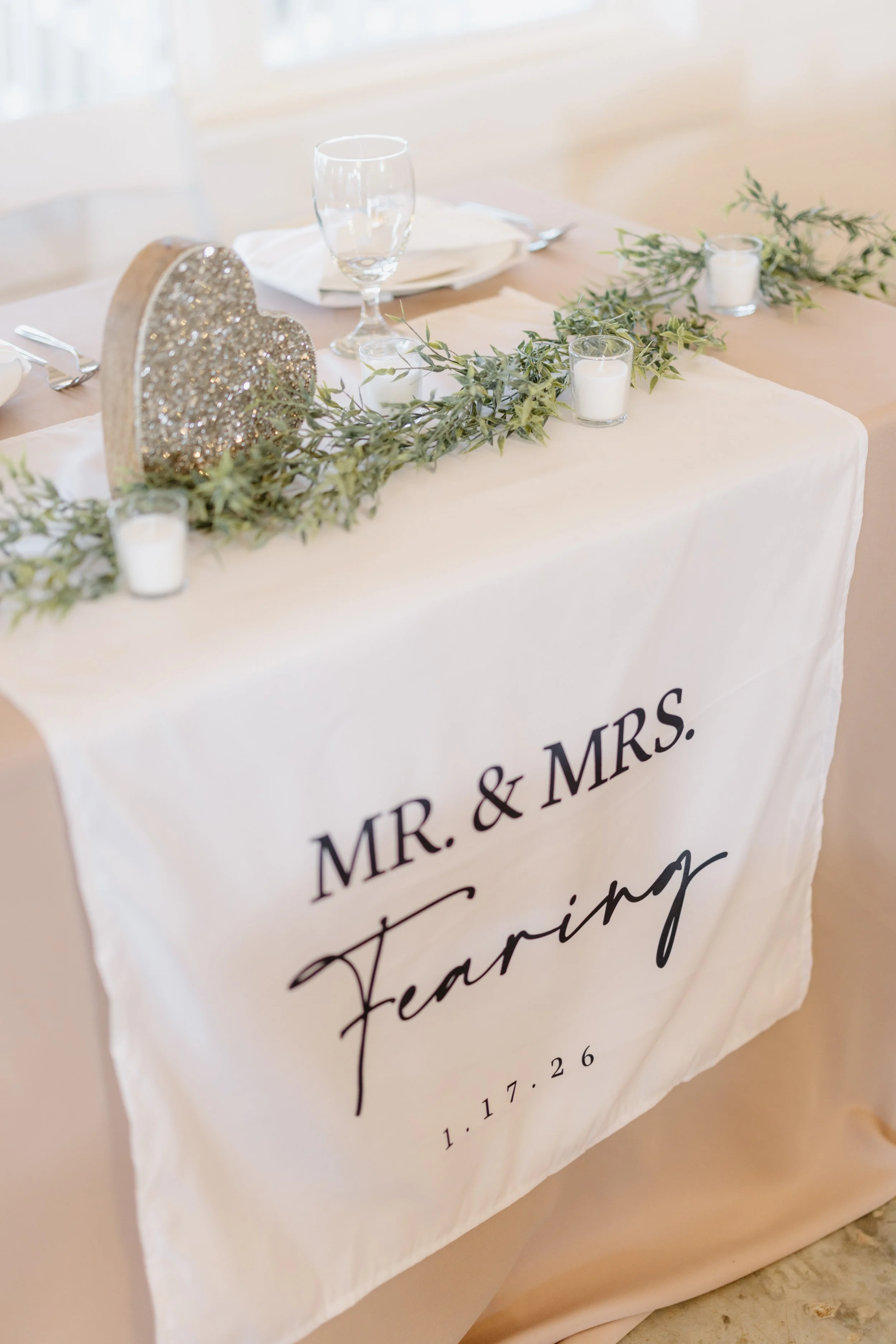 Wedding reception table with a white cloth banner that reads 'Mr. & Mrs. Fearing' and the date '1.17.26,' decorated with a greenery garland, white candles, a glittery heart-shaped decoration, an empty wine glass, and table settings.
