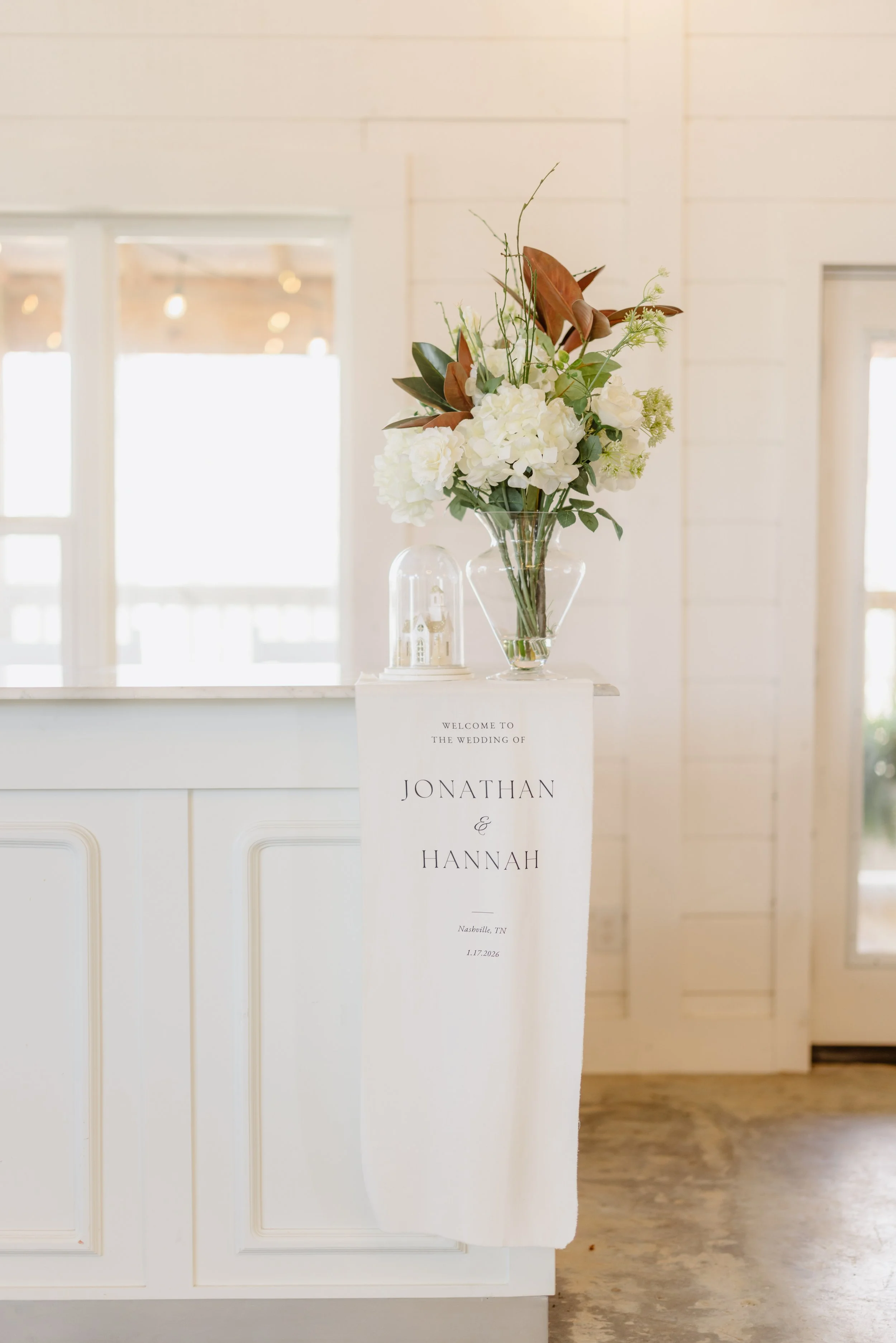 Wedding welcome sign with floral arrangement in glass vase and small decorative building under glass dome on reception counter.