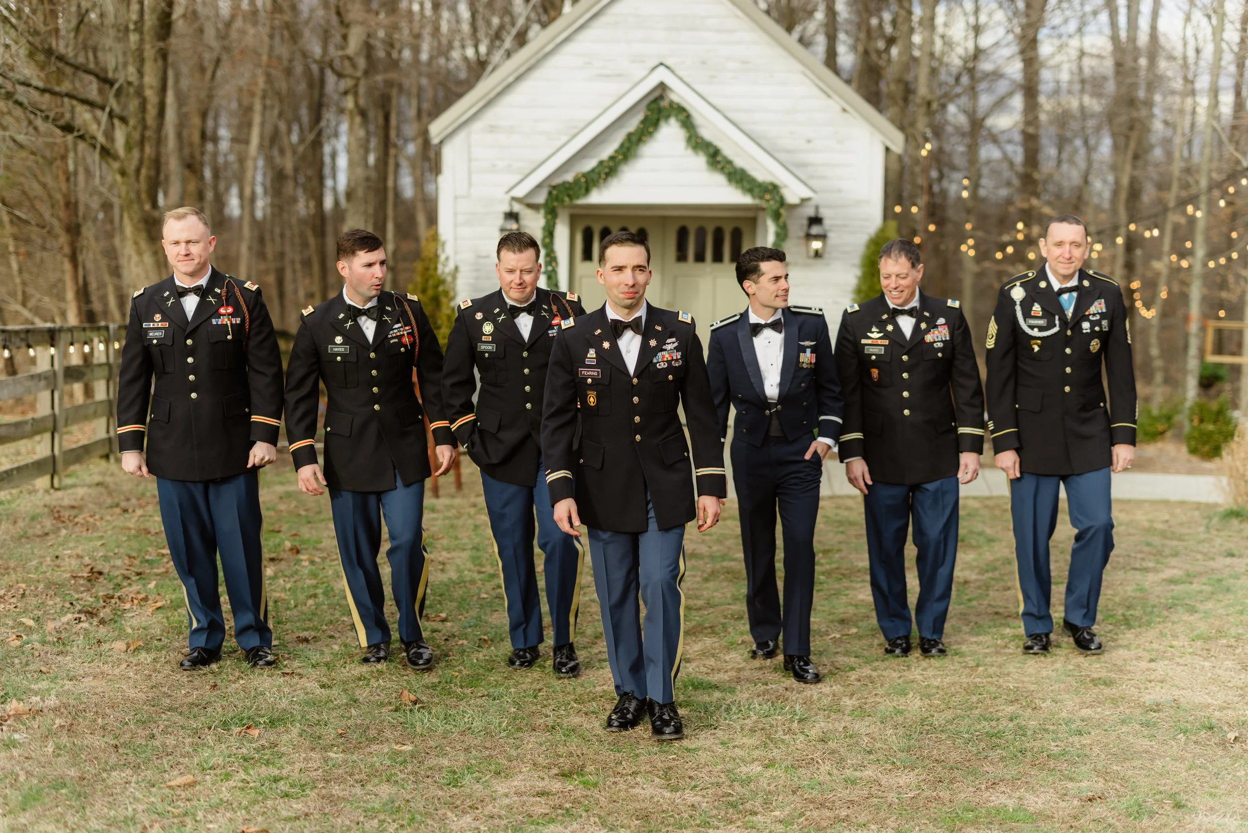 A group of seven men in military formal dress walking outdoors on a grassy area in front of a white building with string lights, trees, and outdoor decorations, possibly at a wedding or special event.