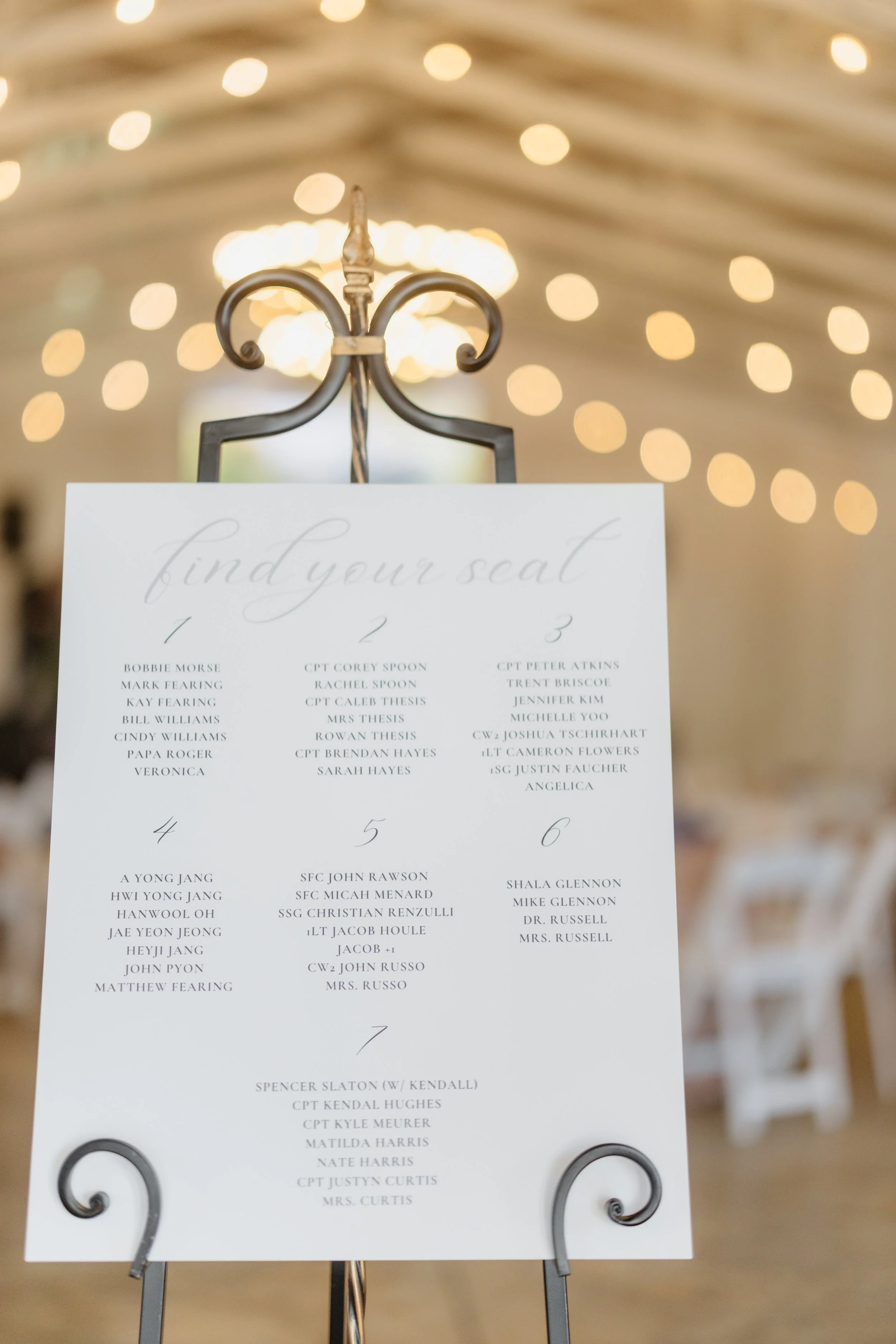 A wedding seating chart with six sections, listing names of guests assigned to each table, displayed on a metal stand in a decorated event hall with hanging string lights and a chandelier in the background.