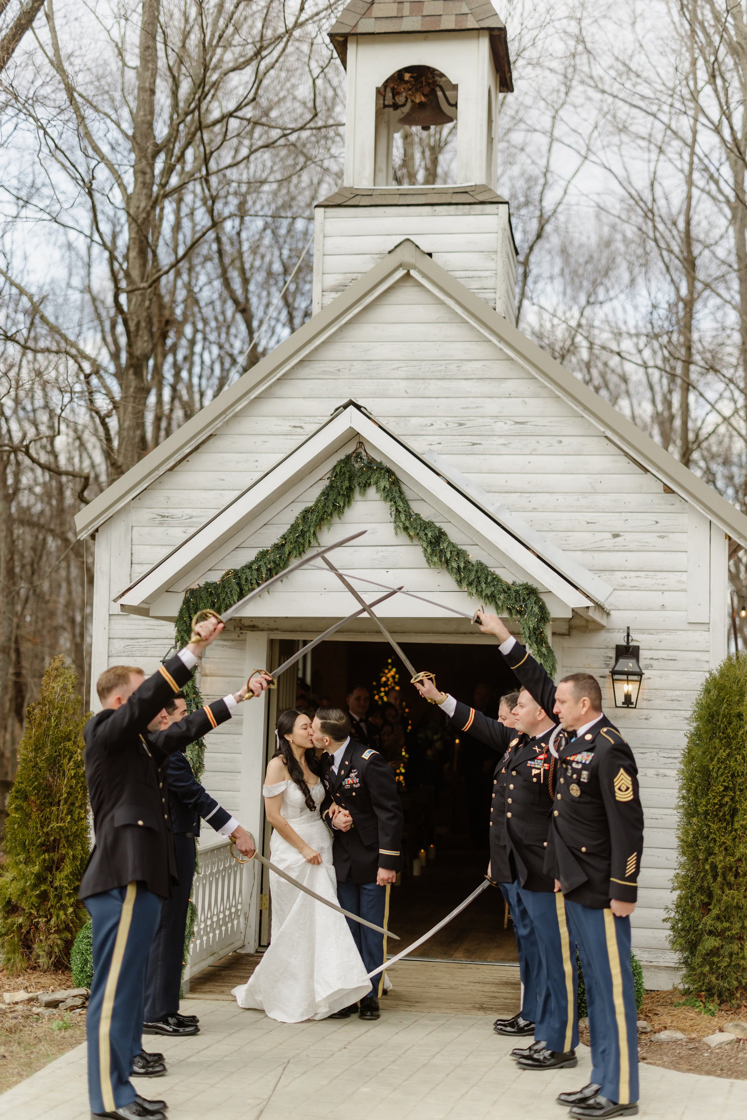 Military wedding with soldiers forming an arch with swords for the bride and groom to kiss outside a small white chapel decorated with greenery.