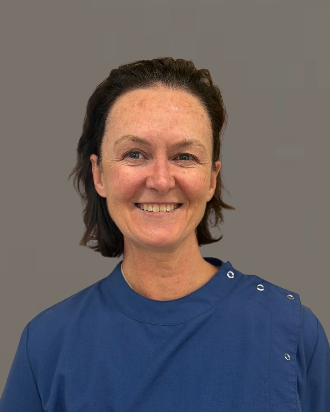 Dr Rebecca Sinclair. Hull. A female Dentist  smiling, wearing a blue dental uniform, standing against a plain background.