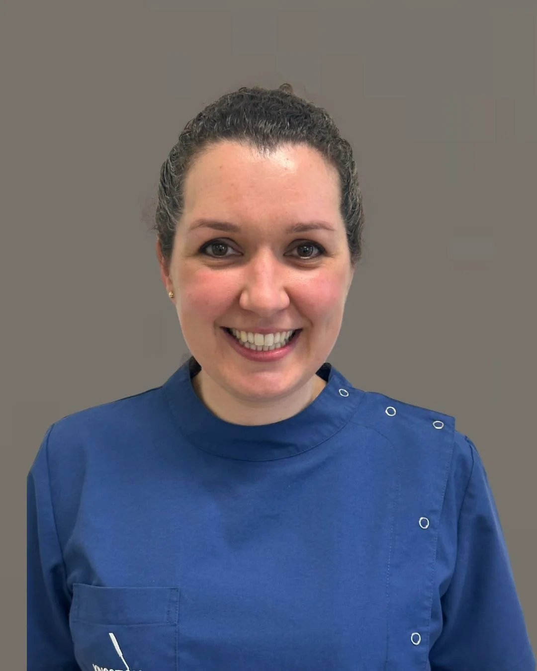 Dr Johanna Hull. A female Dentist with dark curly hair, smiling, wearing a blue dental uniform, standing against a plain background.