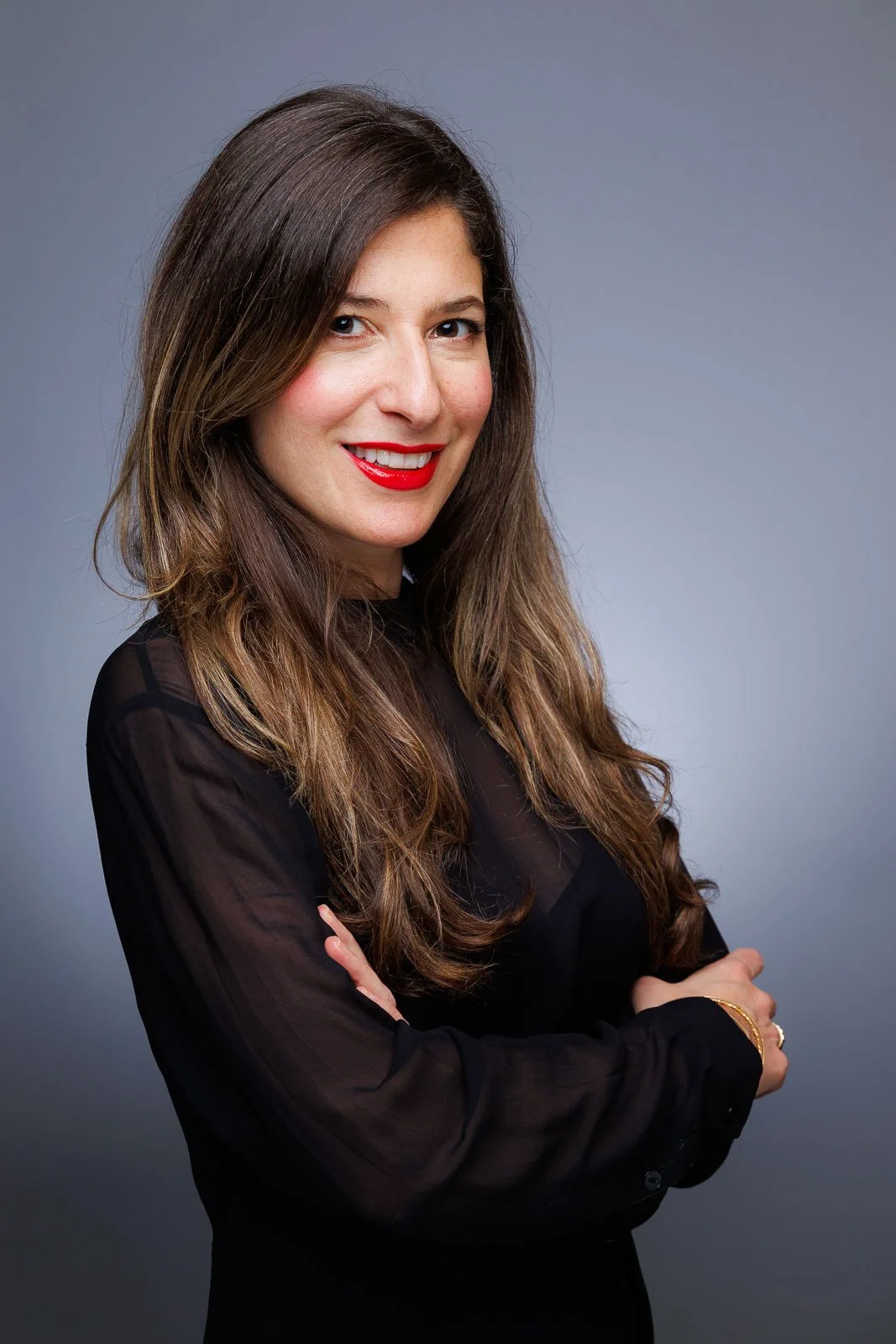 A woman with long, wavy brown hair wearing a black sheer blouse, smiling with red lipstick, crossed arms, against a gray background. User experience design and product designer from New York City