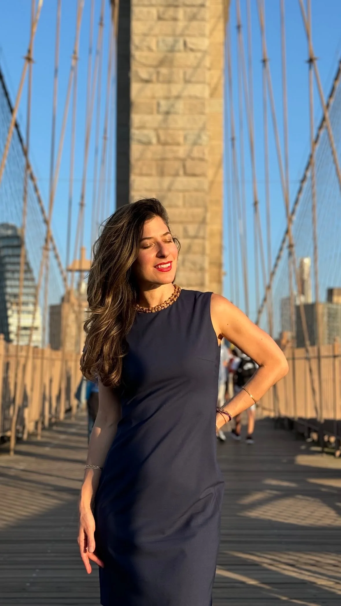 A woman in a navy dress with her eyes closed, standing on a bridge with cables and city buildings in the background, bathed in golden sunlight.