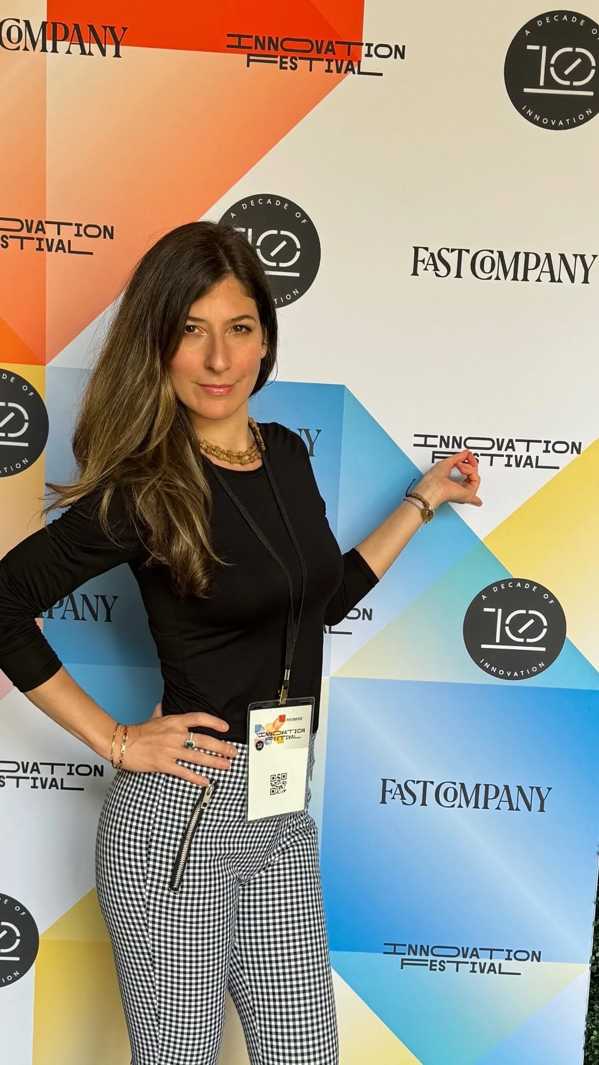 Woman with long brown hair, wearing a black top and black and white checkered pants, standing in front of a colorful backdrop with the words 'Innovation Festival' and other event logos.
