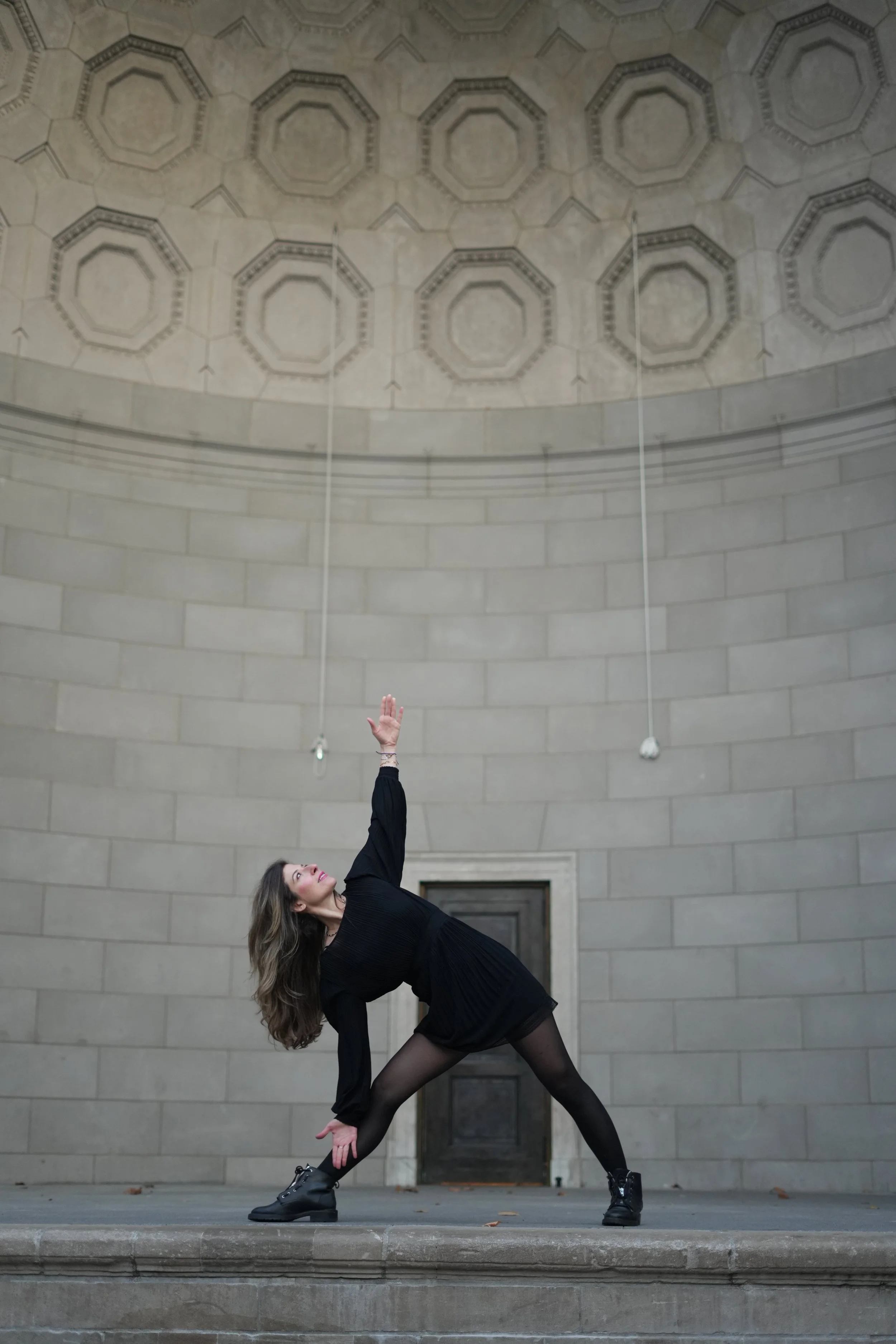 A woman in black dance attire performing a pose on a stone platform in front of a large, curved stone wall with geometric patterns on the ceiling.