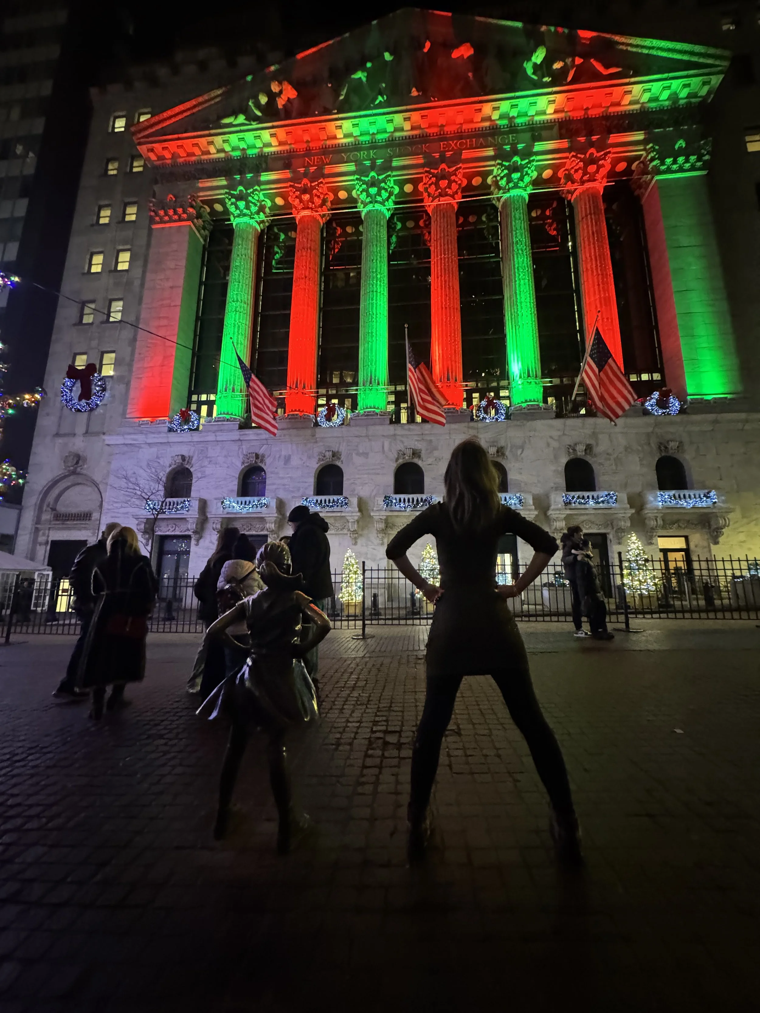 People gathered outside the New York Stock Exchange building decorated with Christmas lights and wreaths, illuminated in red and green for Christmas. User experience design and product designer from New York City