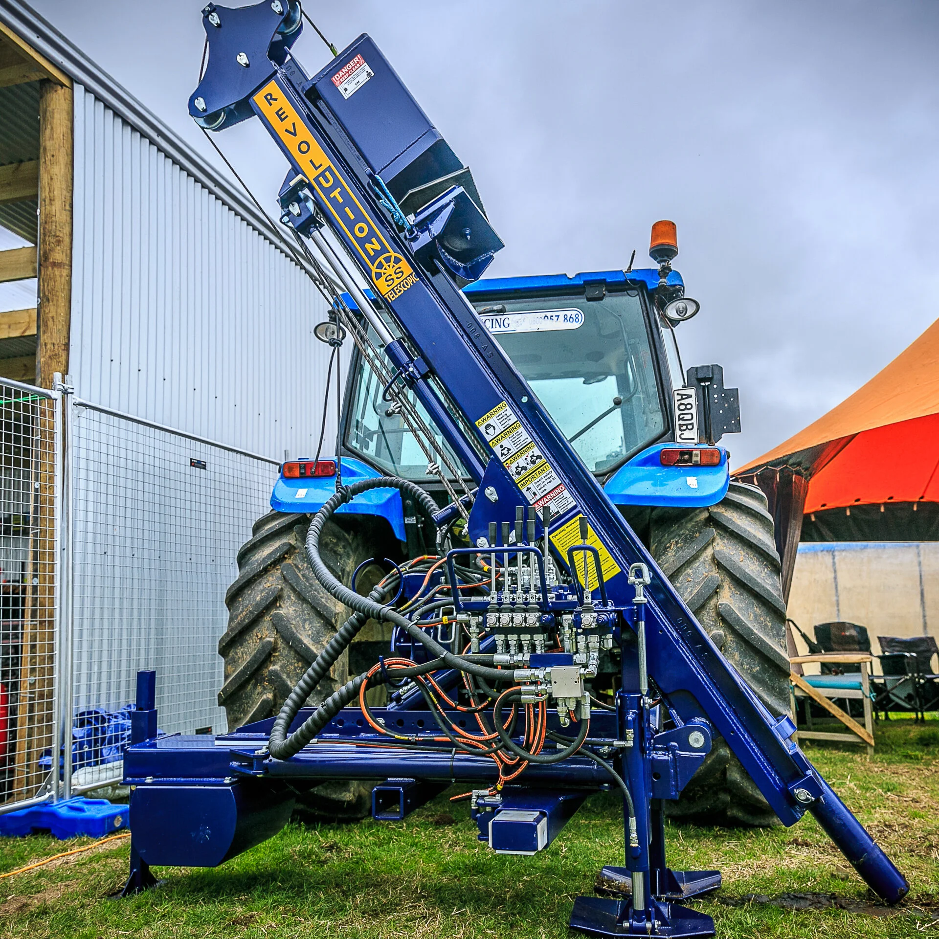 A large tractor with a hydraulic post driver, parked outdoors on grass with a building structure and cloudy sky in the background.