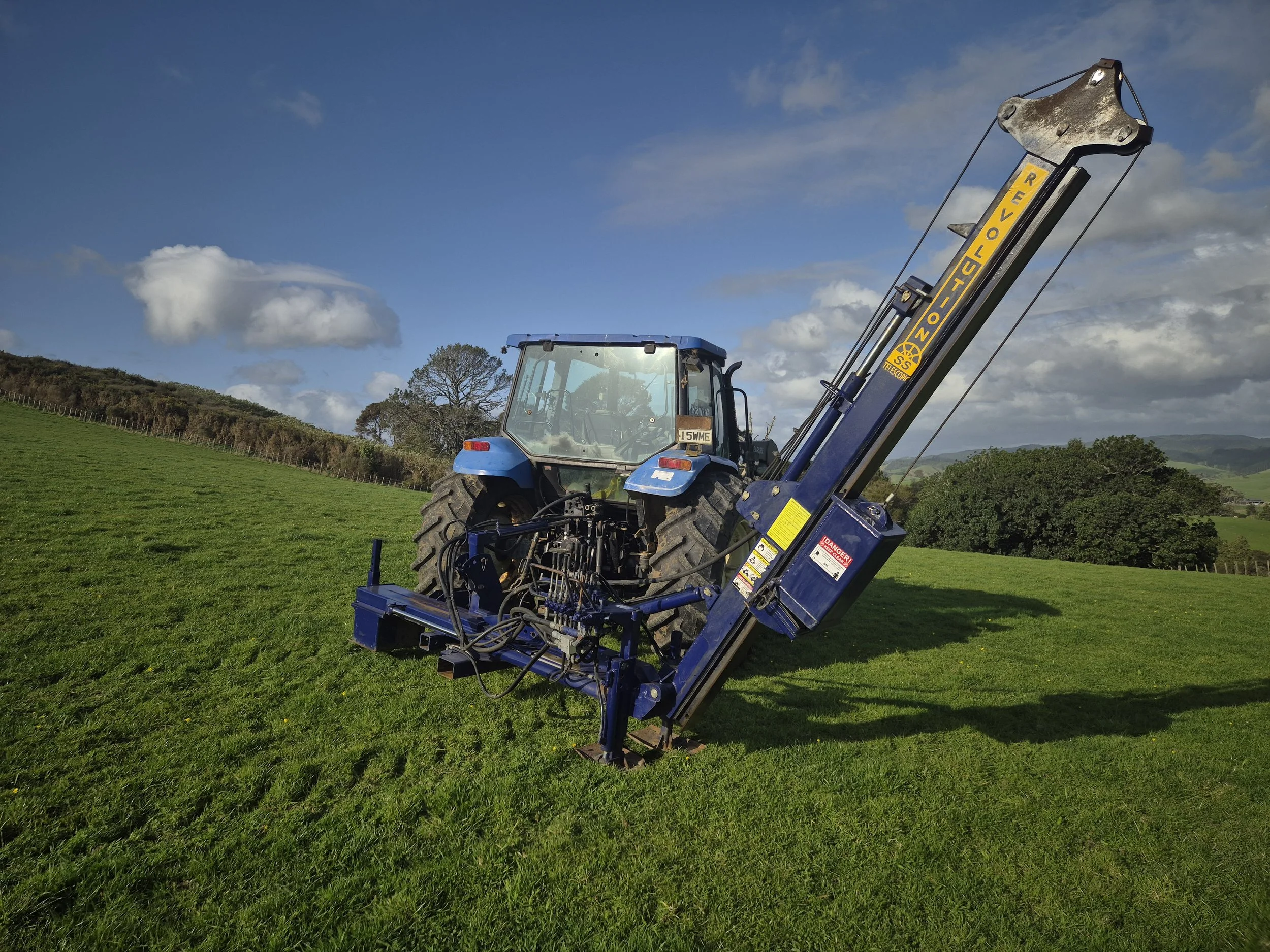 A blue tractor with a mounted post rammer in a green field under a partly cloudy sky.