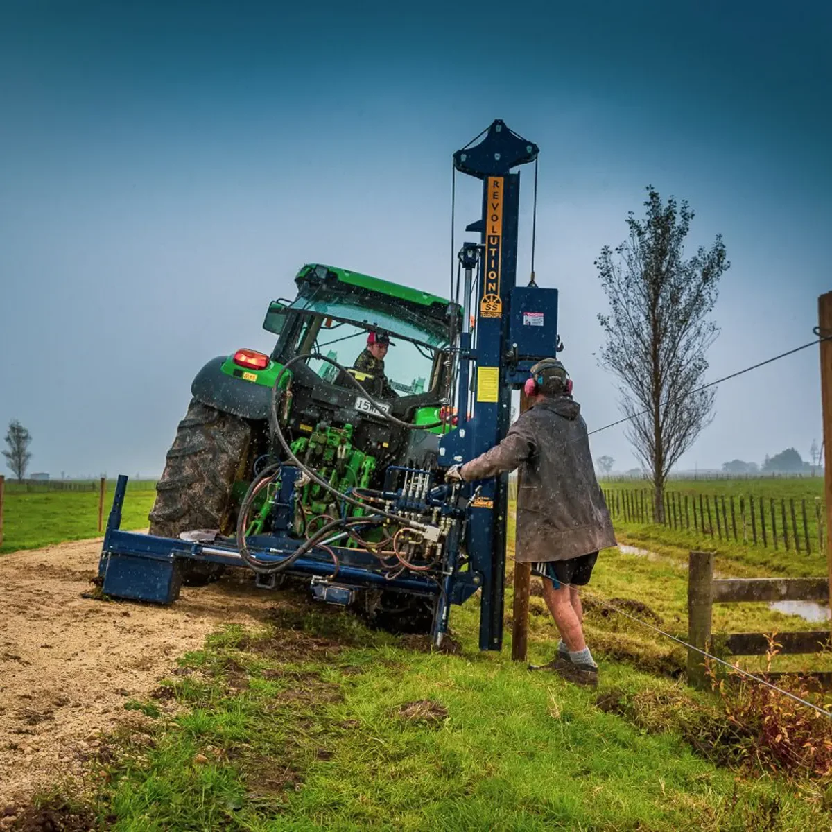 A person operating a tractor with a large post ramming rig attachment in a field during rainy weather.