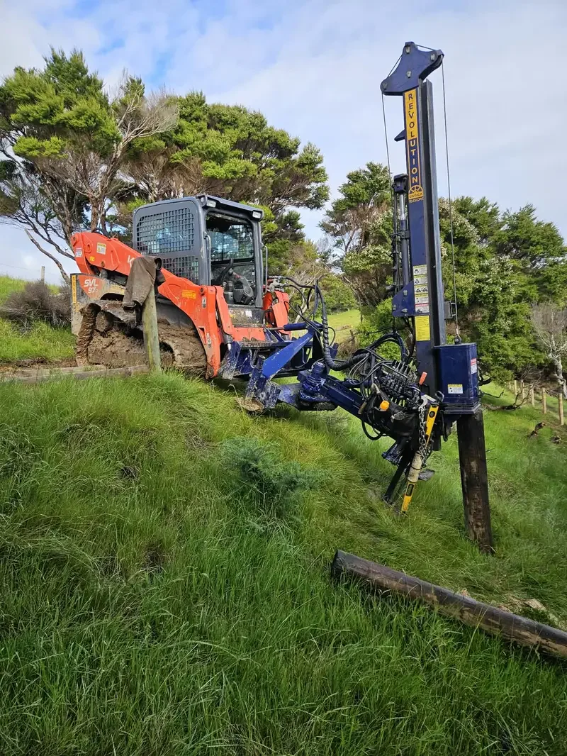 A construction vehicle, specifically a tracked excavator or skid-steer loader, is tilted with part of its body over a grassy hillside and a fencing post driver attached to it driving in a fence post.
