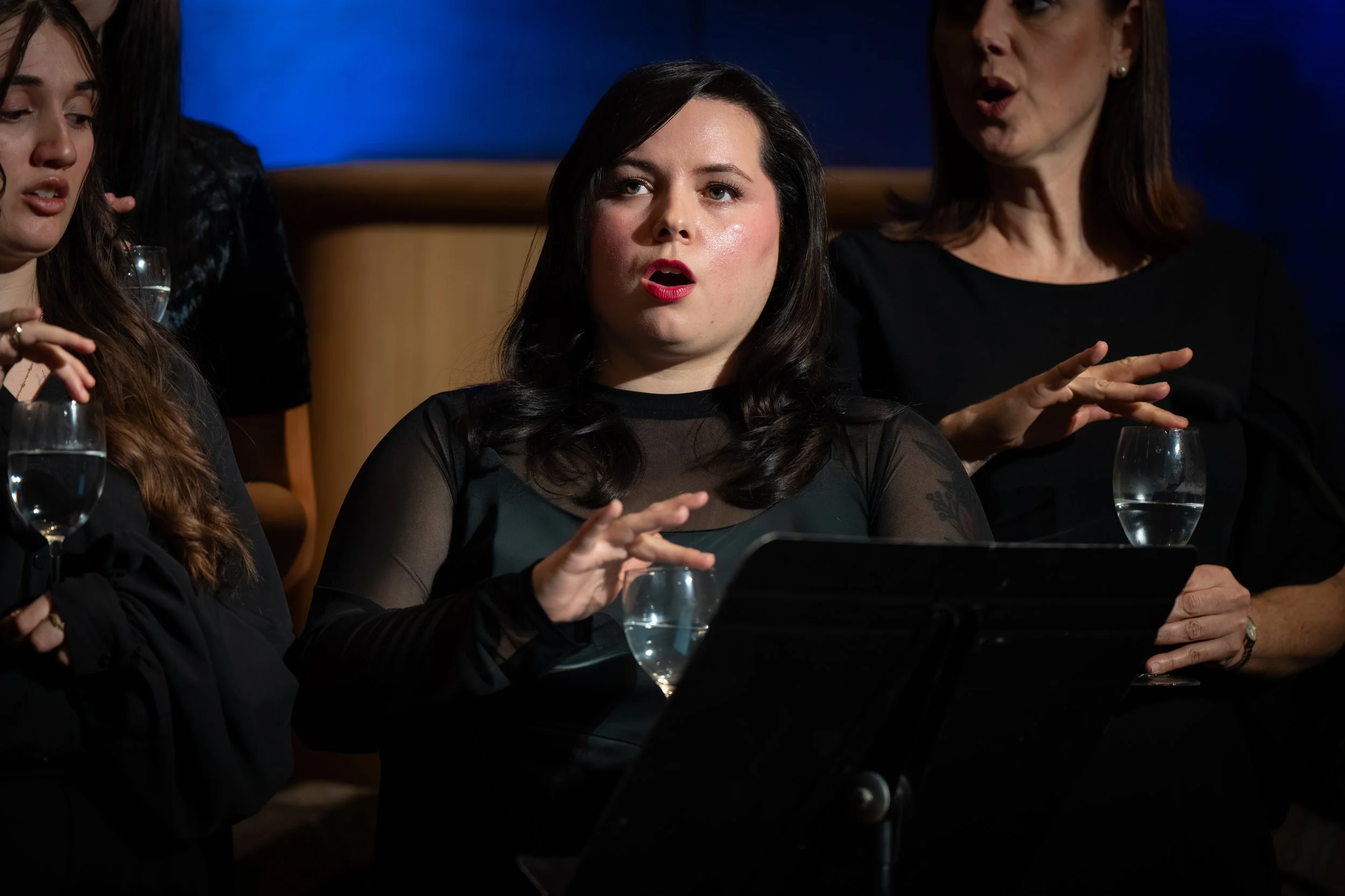 Chorus singers perform with water glasses, focusing intently during a musical piece with dramatic lighting.