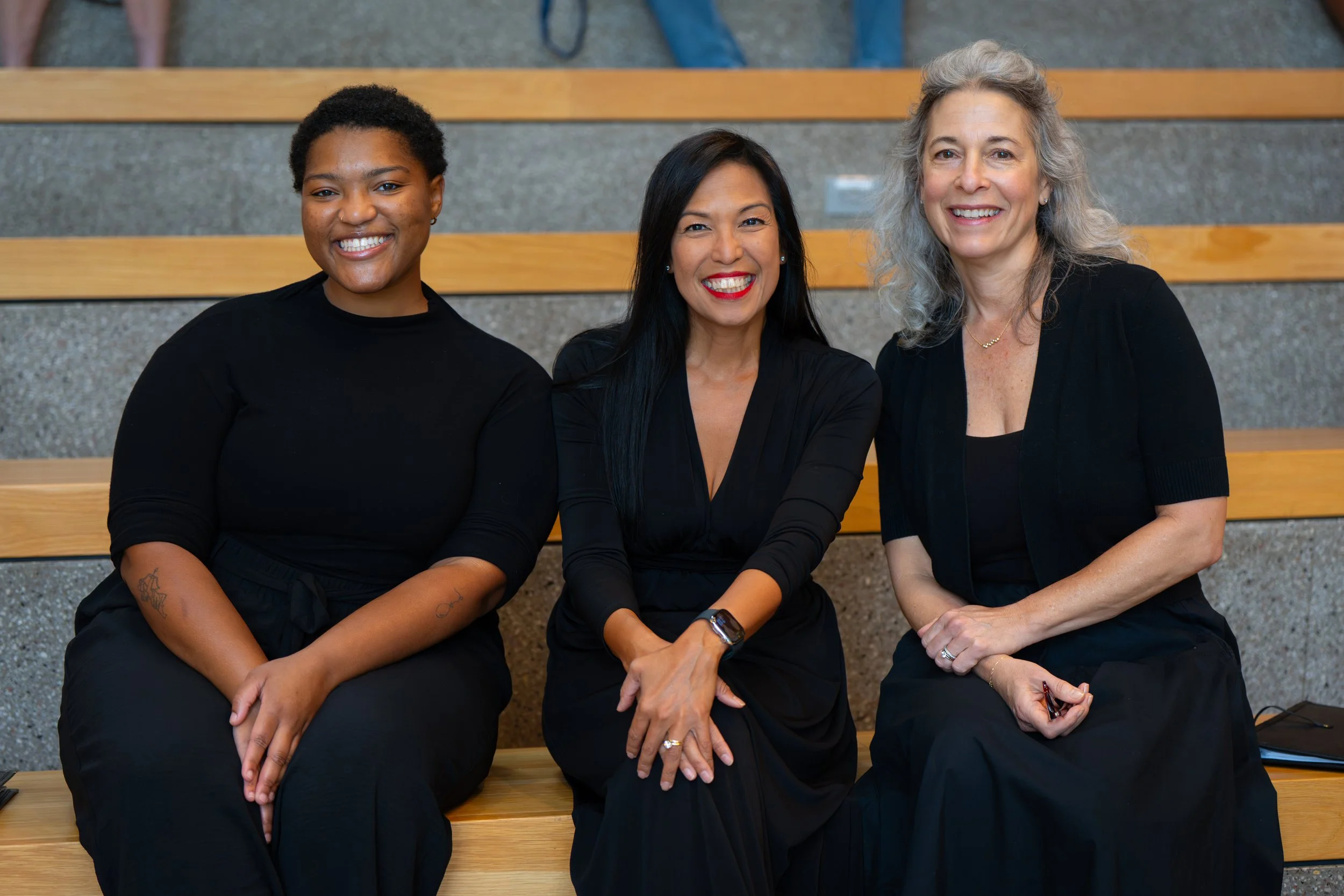 Three singers in black concert attire sit on wooden steps, smiling warmly.