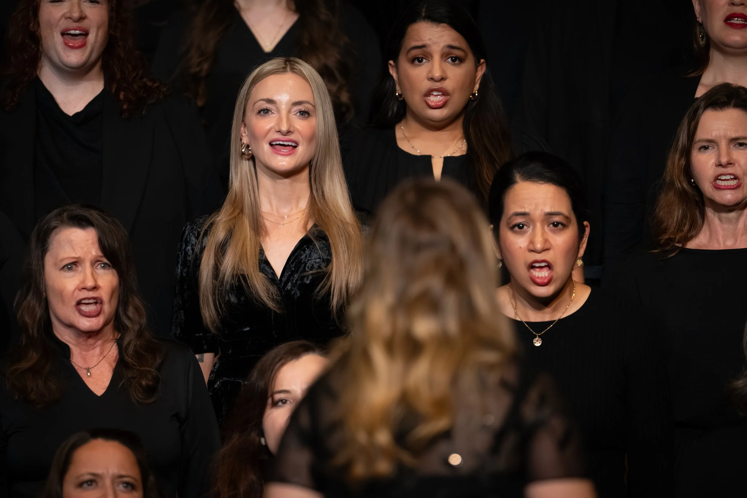 Chorus members sing expressively during a performance, facing their conductor in coordinated black attire.