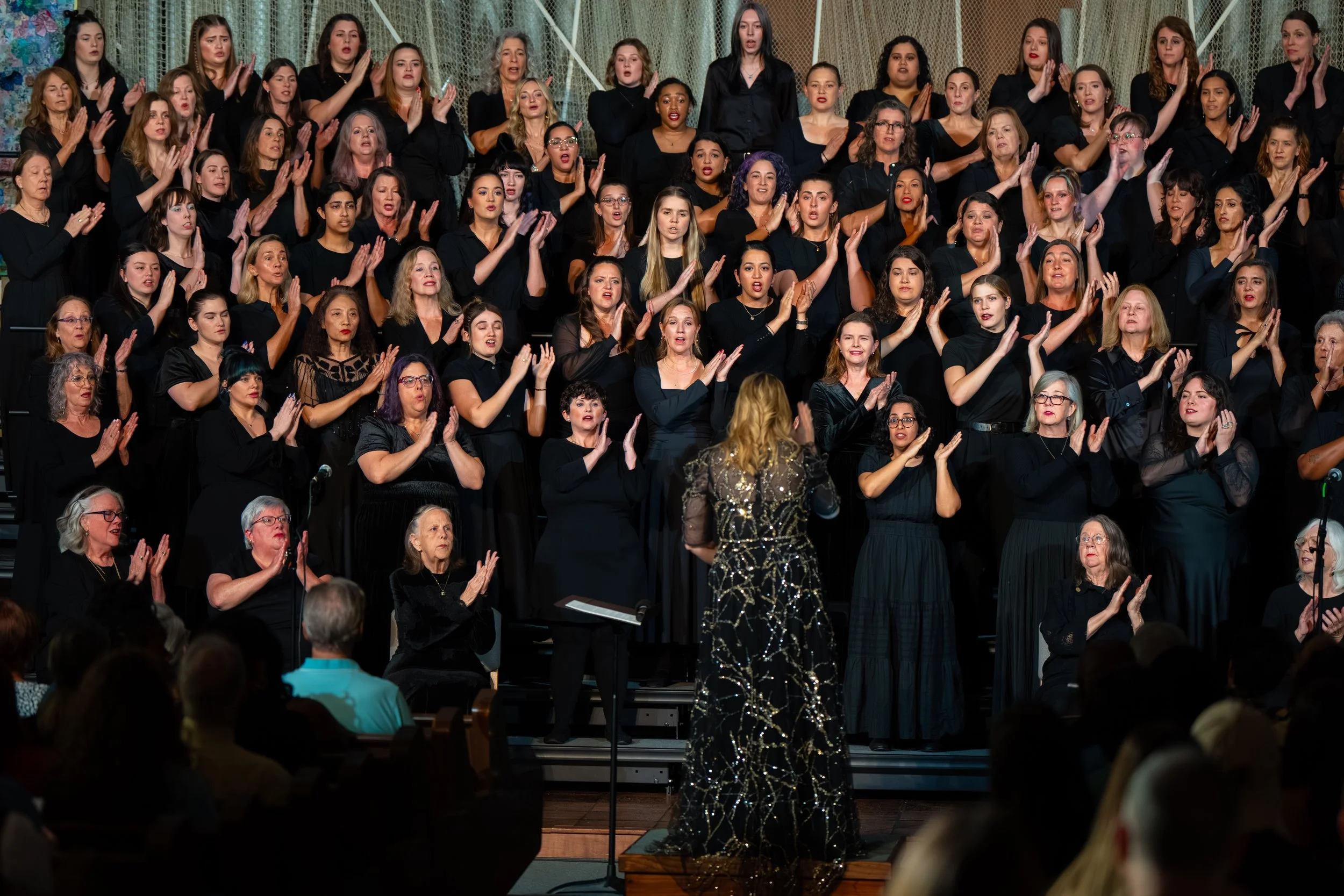 Large choir in black singing and clapping in rhythm while their conductor leads from the front of the stage.
