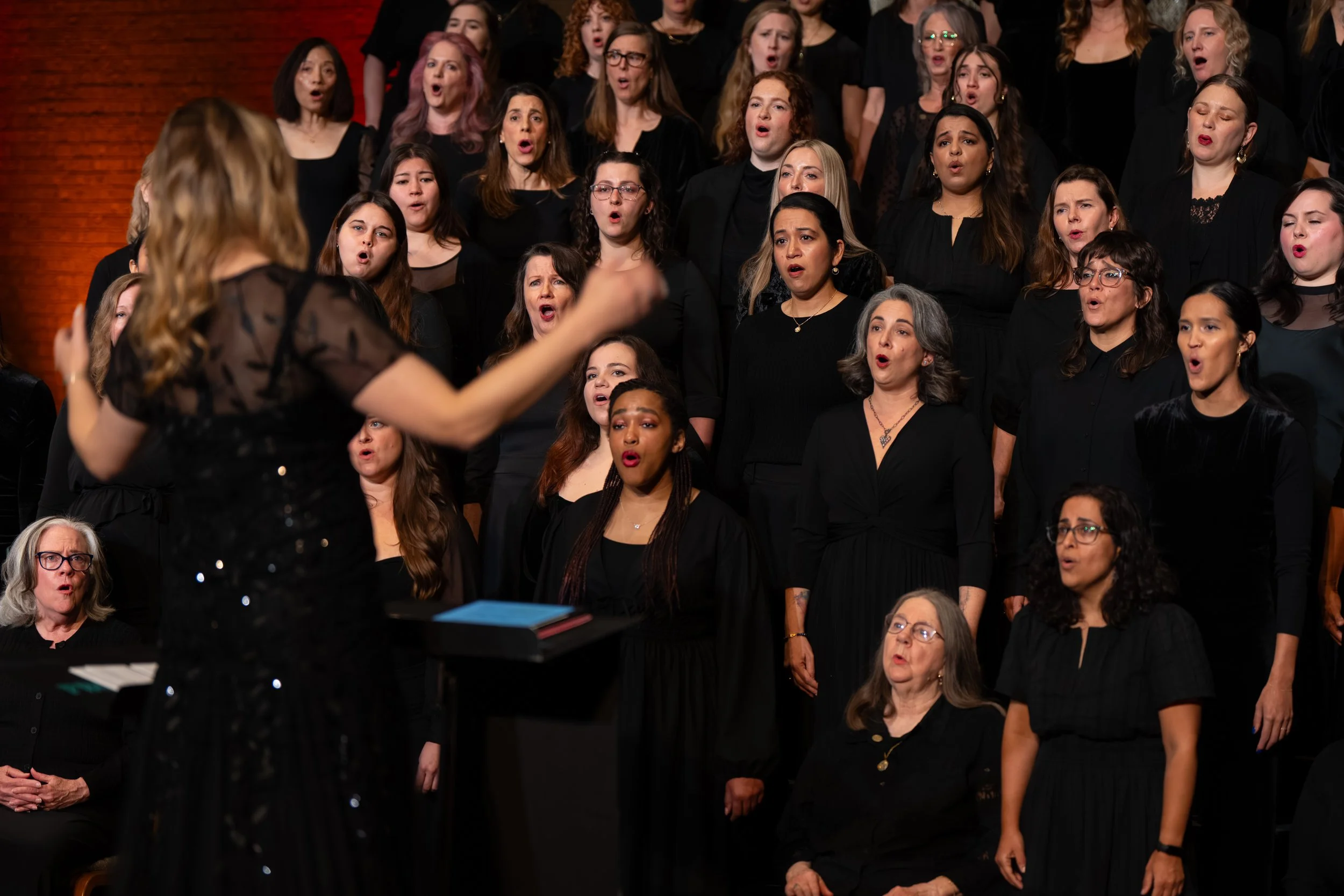 Large treble chorus performs on stage in black outfits, led by conductor with arms raised mid-gesture.