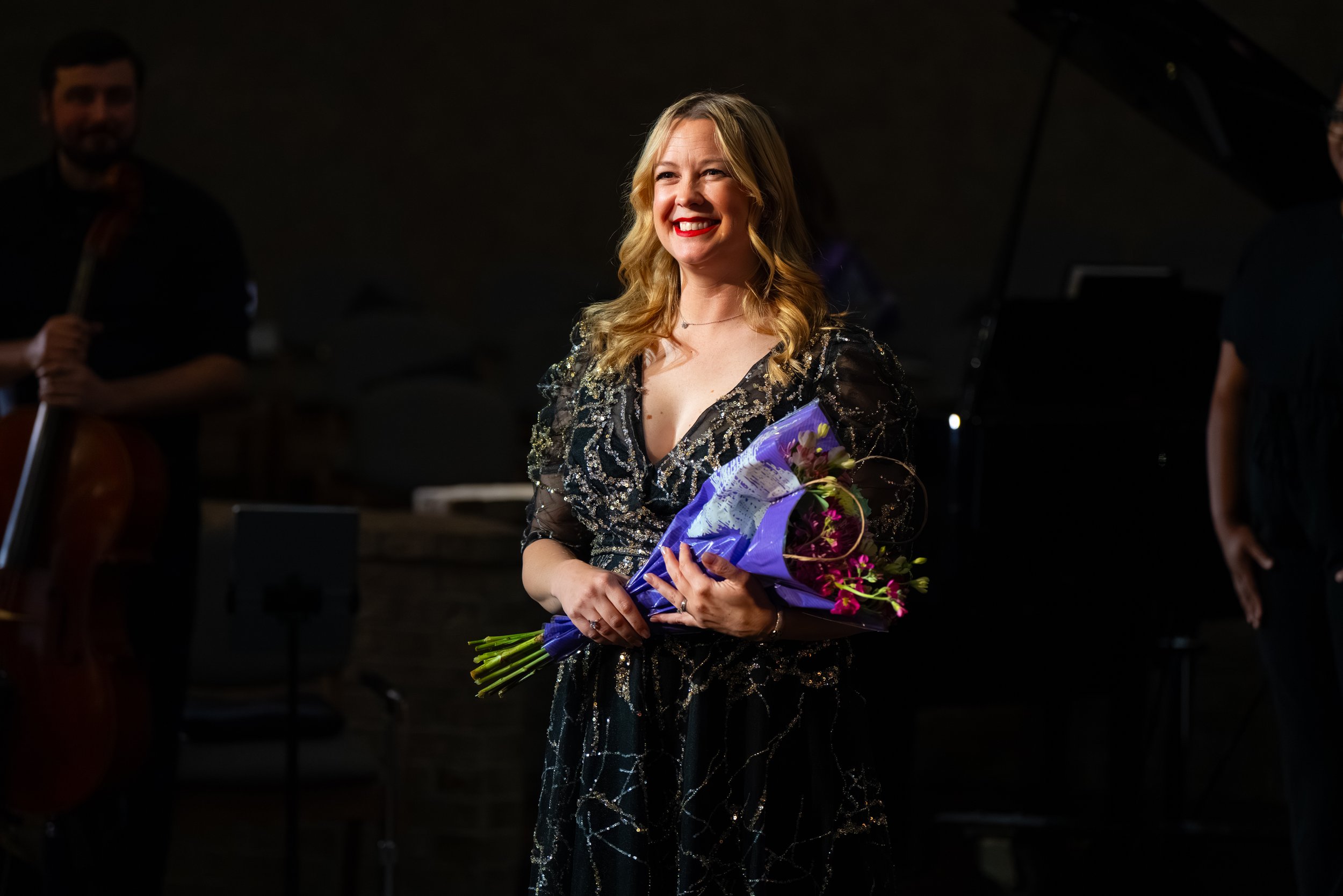 Artistic Director Lisa Sunset Holt in a sequined black gown smiling and holding a bouquet of flowers on stage after a performance.