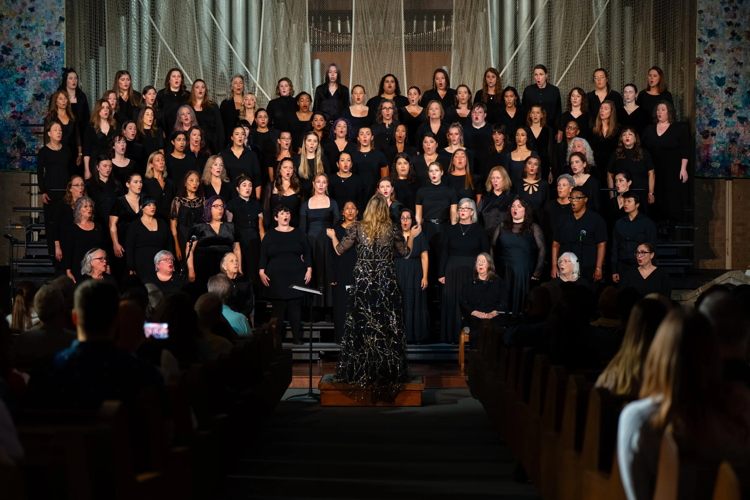 Women's choir singing on risers, seen from the audience with the conductor directing from the podium.