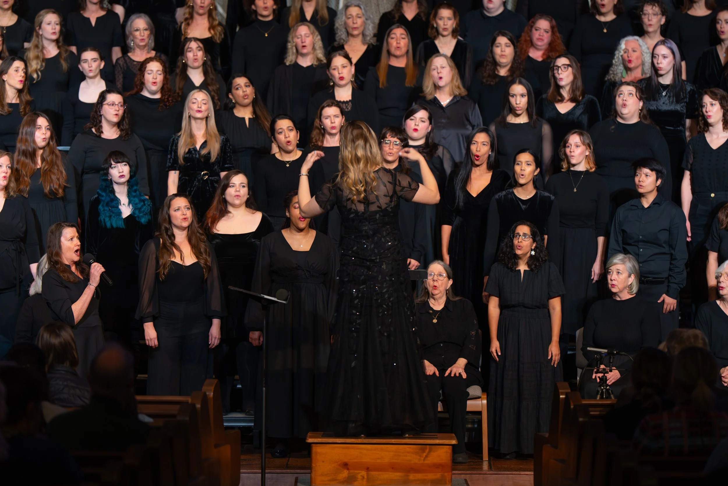 Women's choir performing in black formal attire with conductor leading singers during concert performance