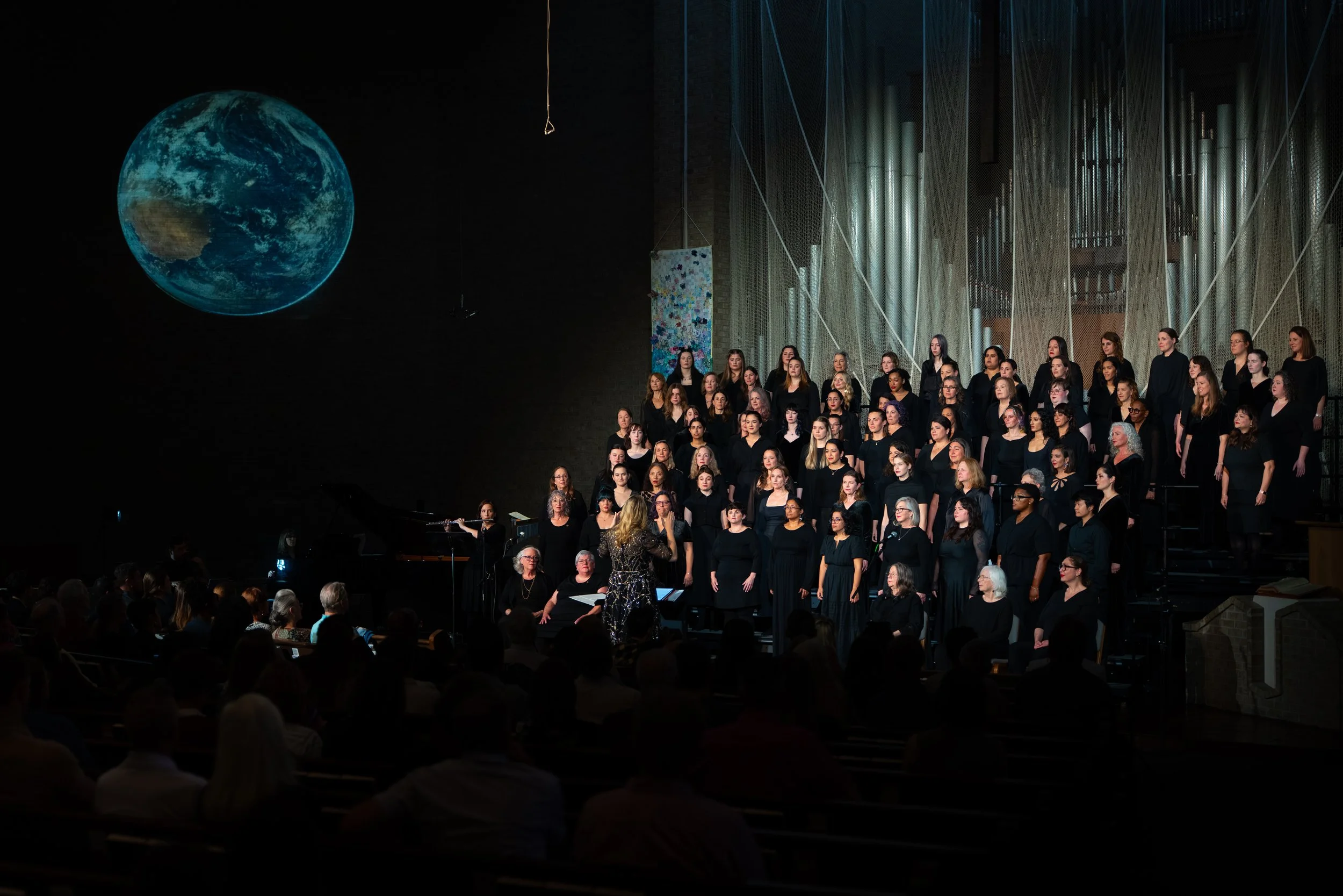 Wide view of a choir performing on risers beneath a large projected image of Earth, with conductor, flutist, and pianist in front of a full audience.