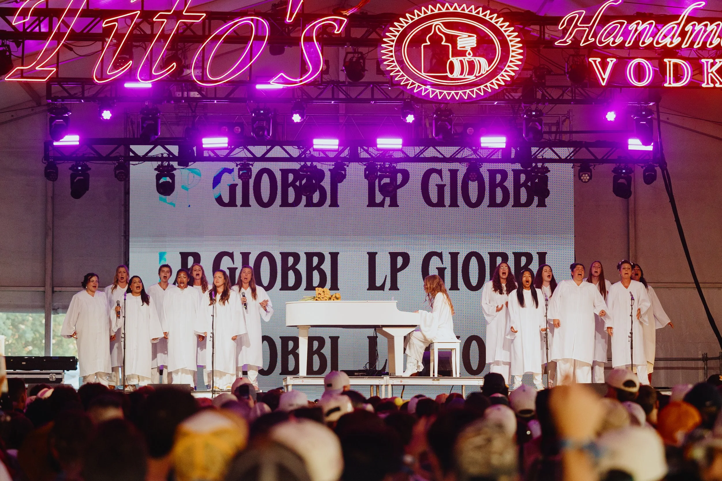 Community chorus in white robes sings on the ACL Festival stage beside a white piano, performing for a large outdoor crowd