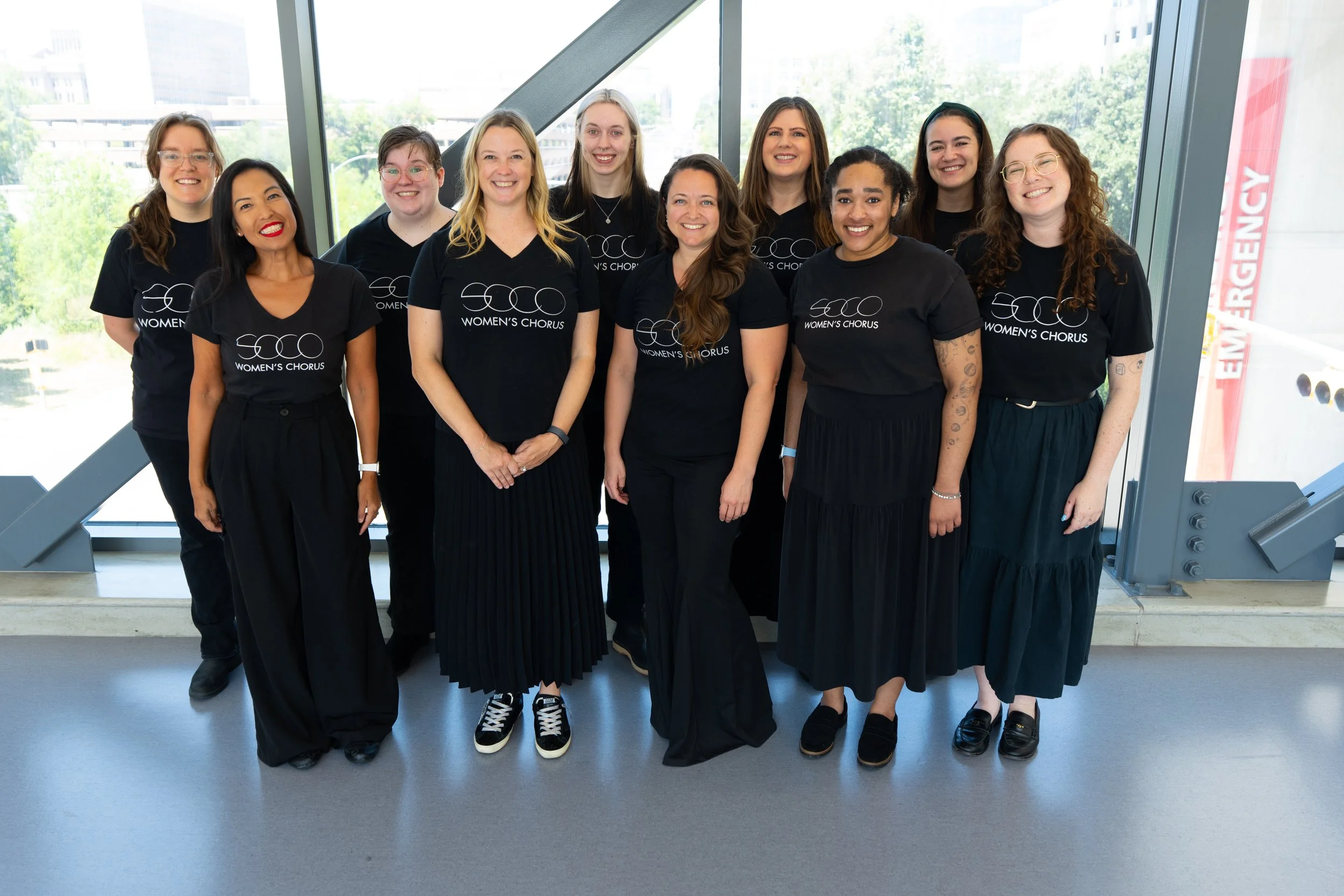 Ten SoCo Women’s Chorus members pose together in black chorus attire inside a bright, window-lined building.