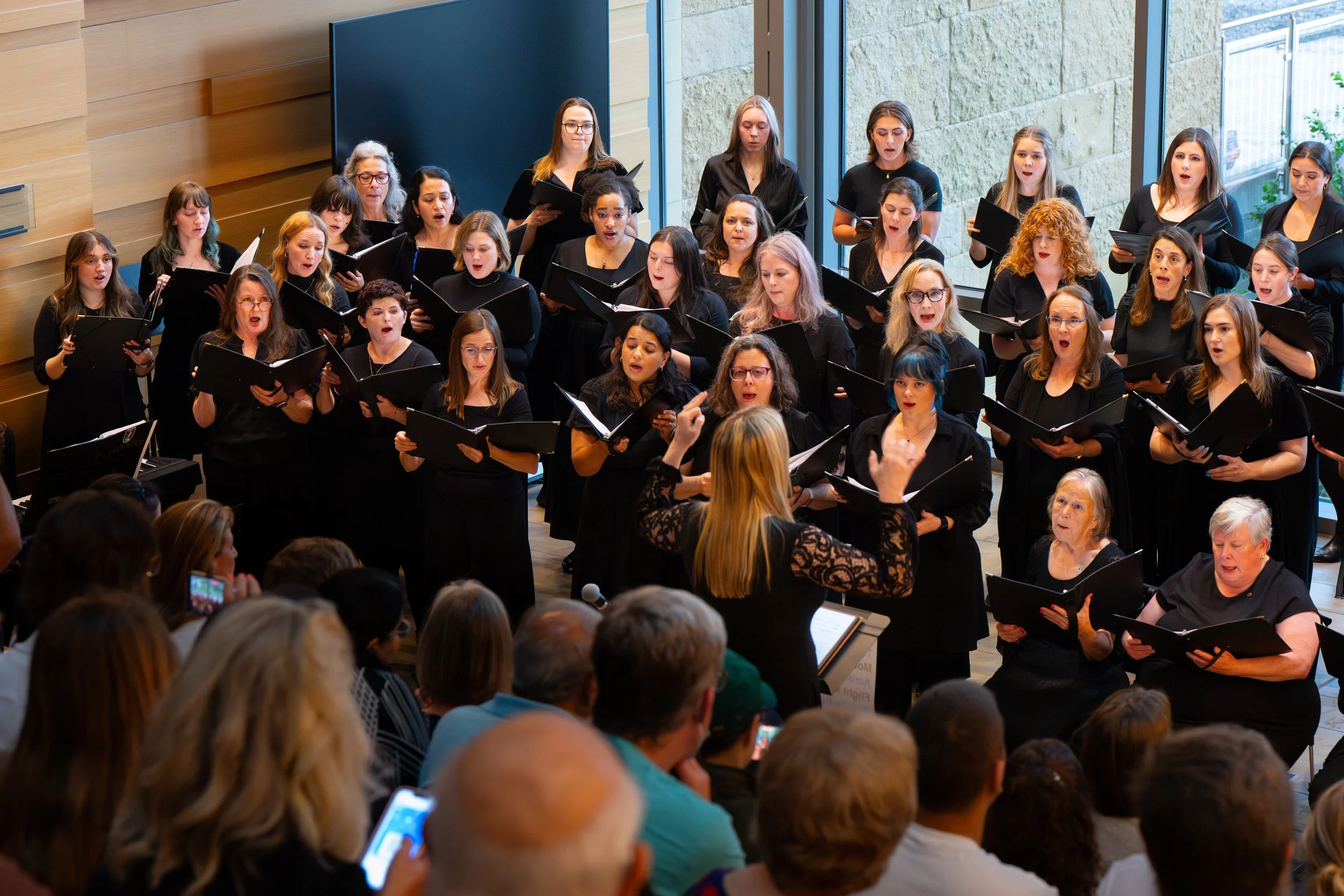 Large treble chorus performs on stage in black outfits, led by conductor with arms raised mid-gesture.