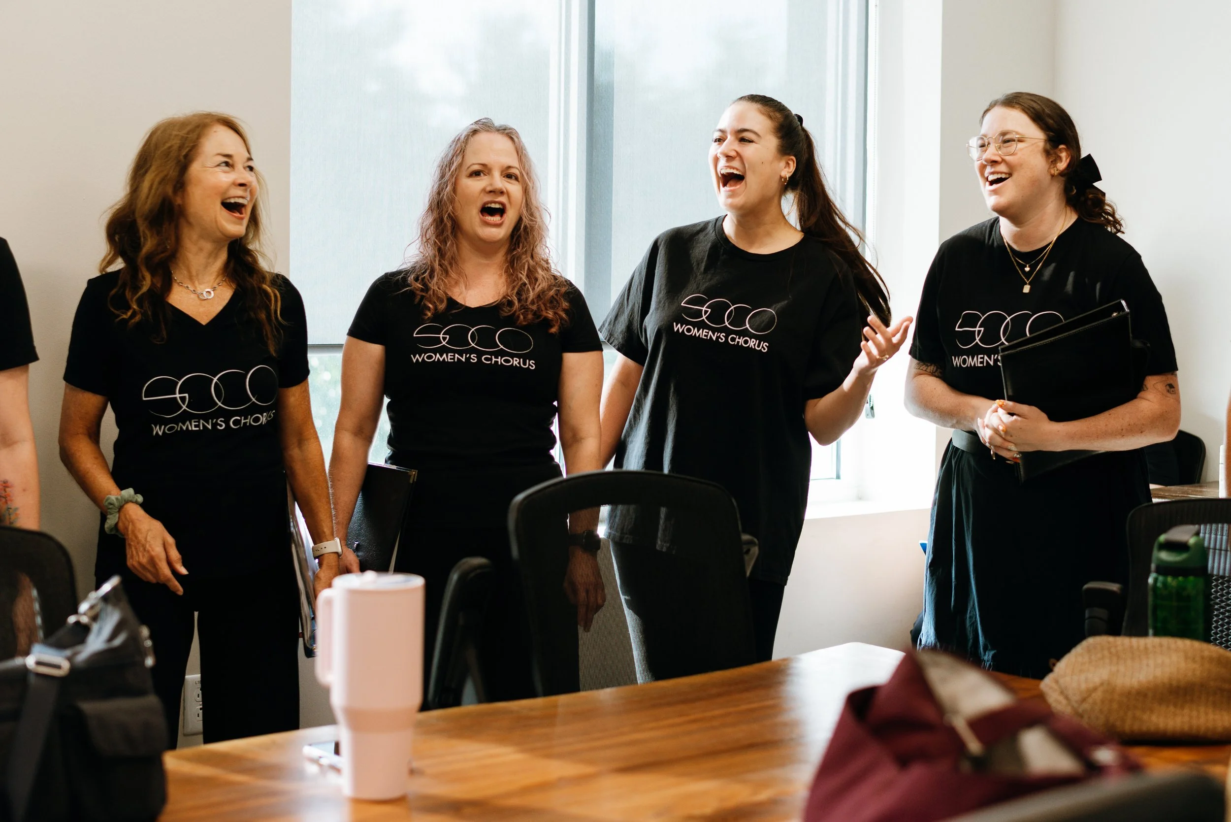 Four SoCo Women’s Chorus members sing together during a rehearsal or informal performance in a sunlit room.