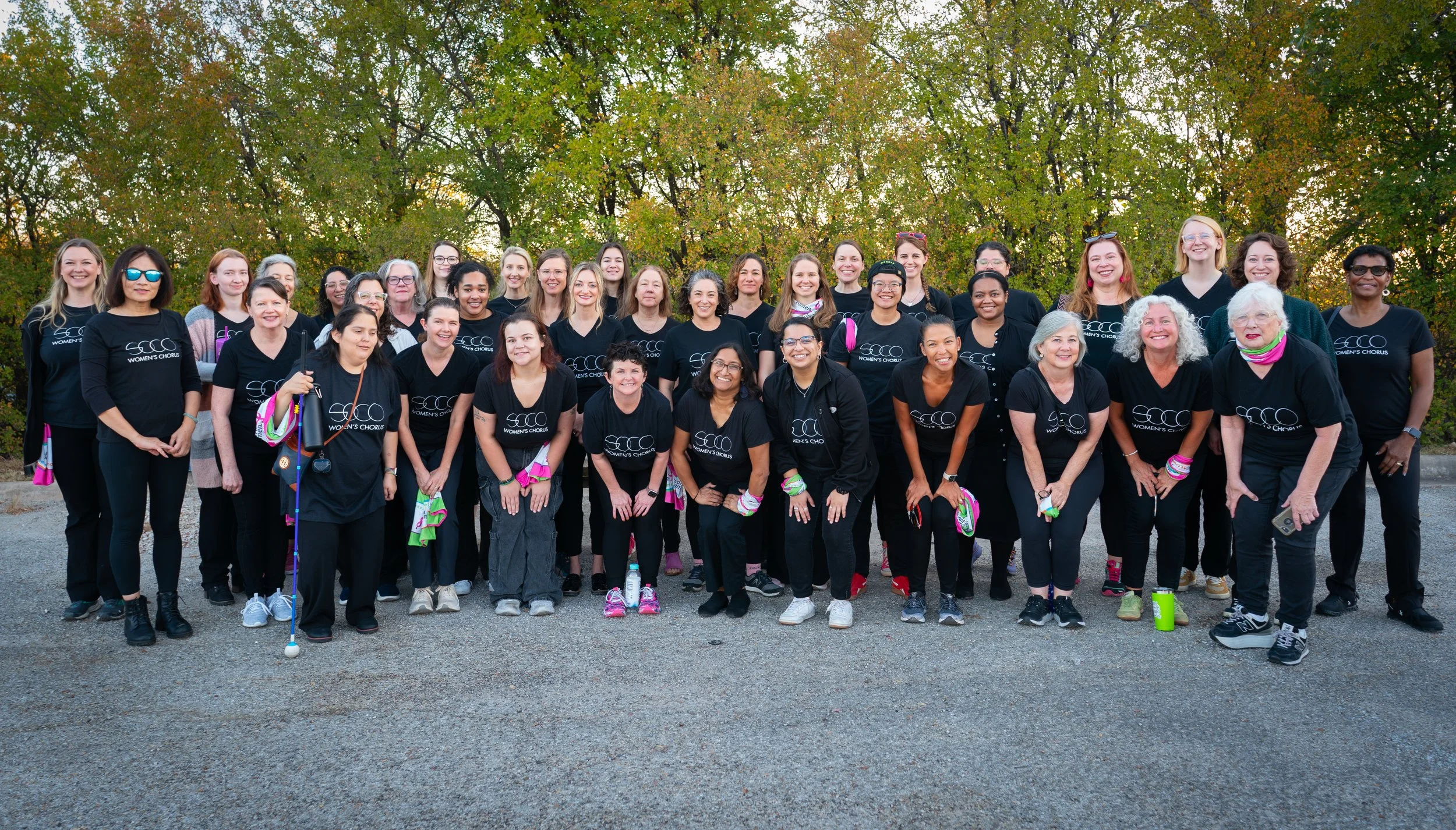 Large, diverse group of women from a community chorus stand together outdoors in matching black shirts, smiling after a performance