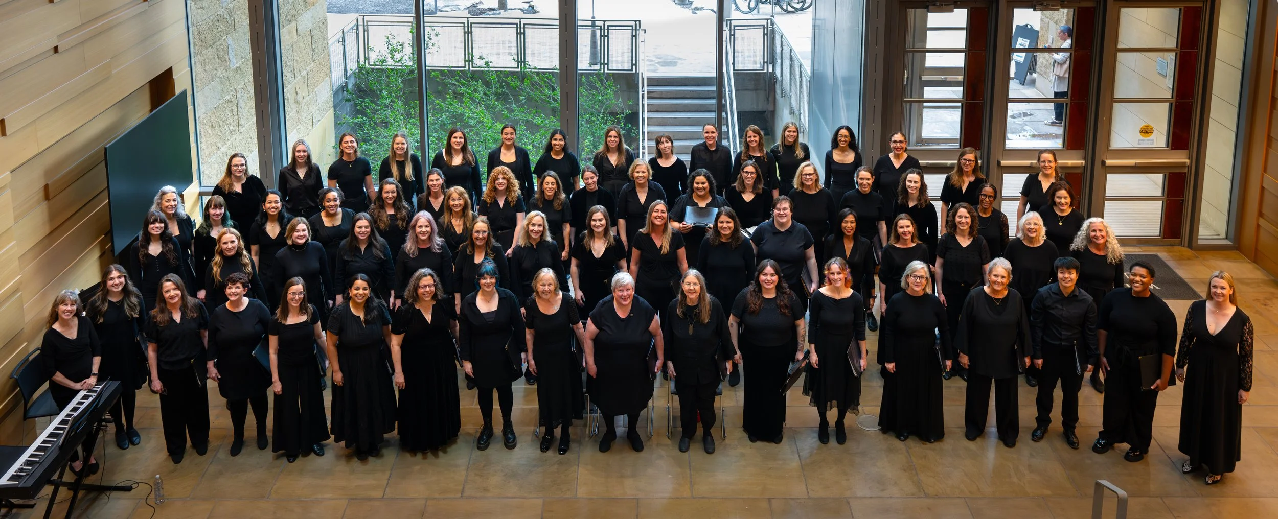 Large community chorus of diverse singers in black attire stands together in a bright atrium, smiling and ready to perform, showcasing inclusivity, music, and group connection.