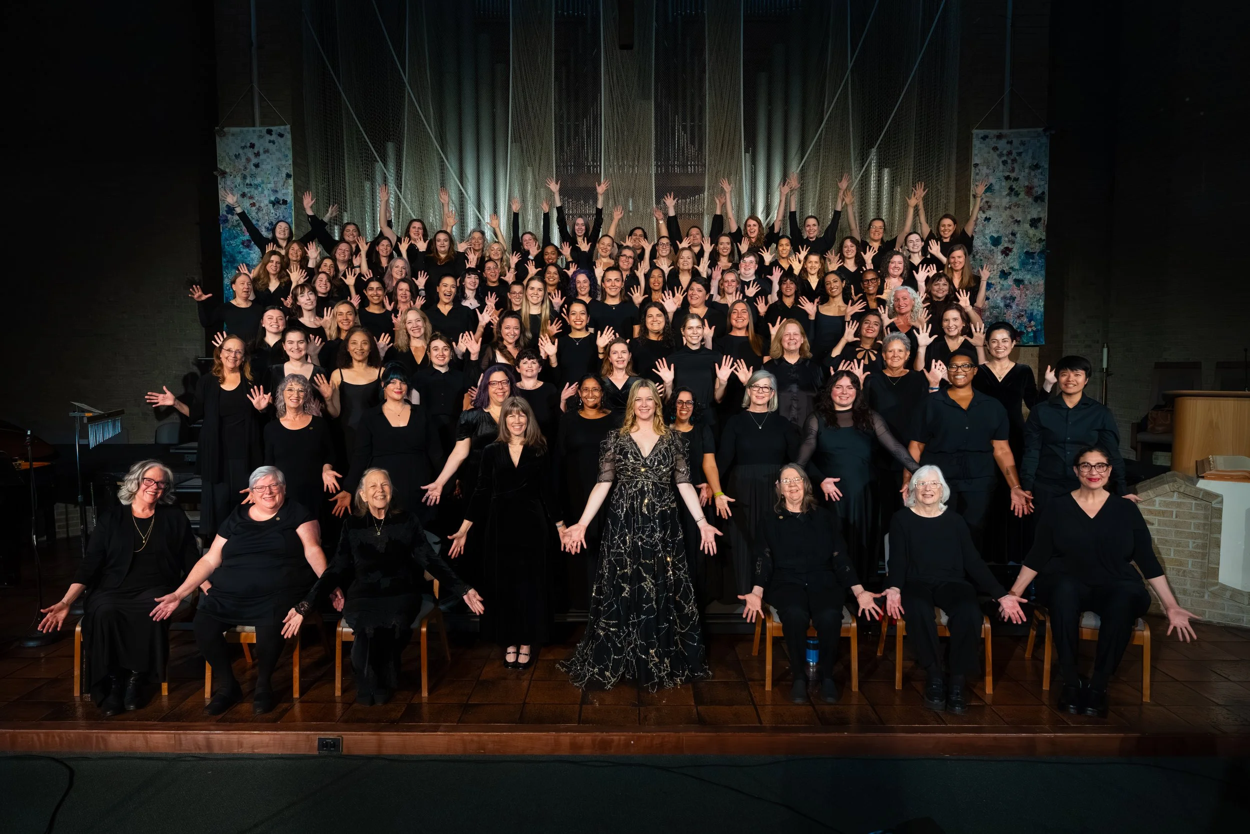 Full women's choir group portrait on stage in black attire, hands raised and outstretched, with the conductor standing center front.