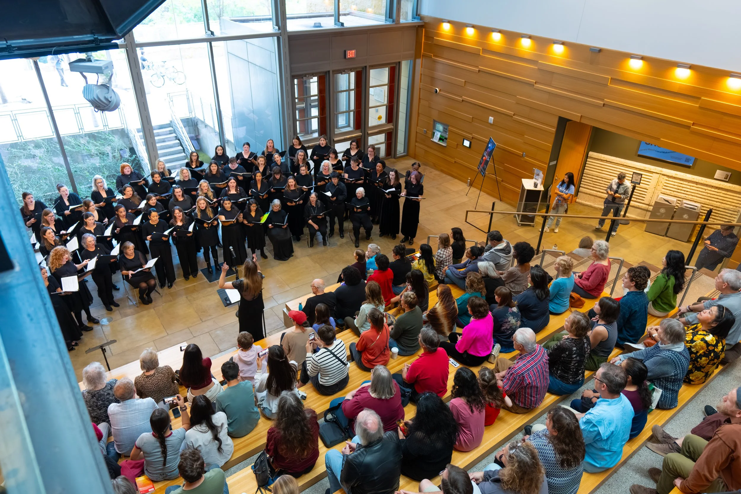 SoCo Women’s Chorus performs in black attire for a seated audience at Austin Central Library atrium.
