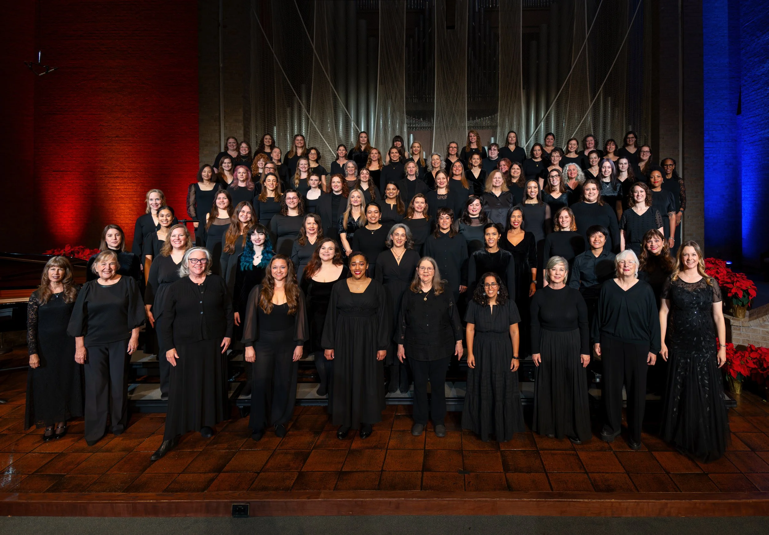 Full community chorus posed on risers in formal black attire, smiling in a warmly lit concert hall.