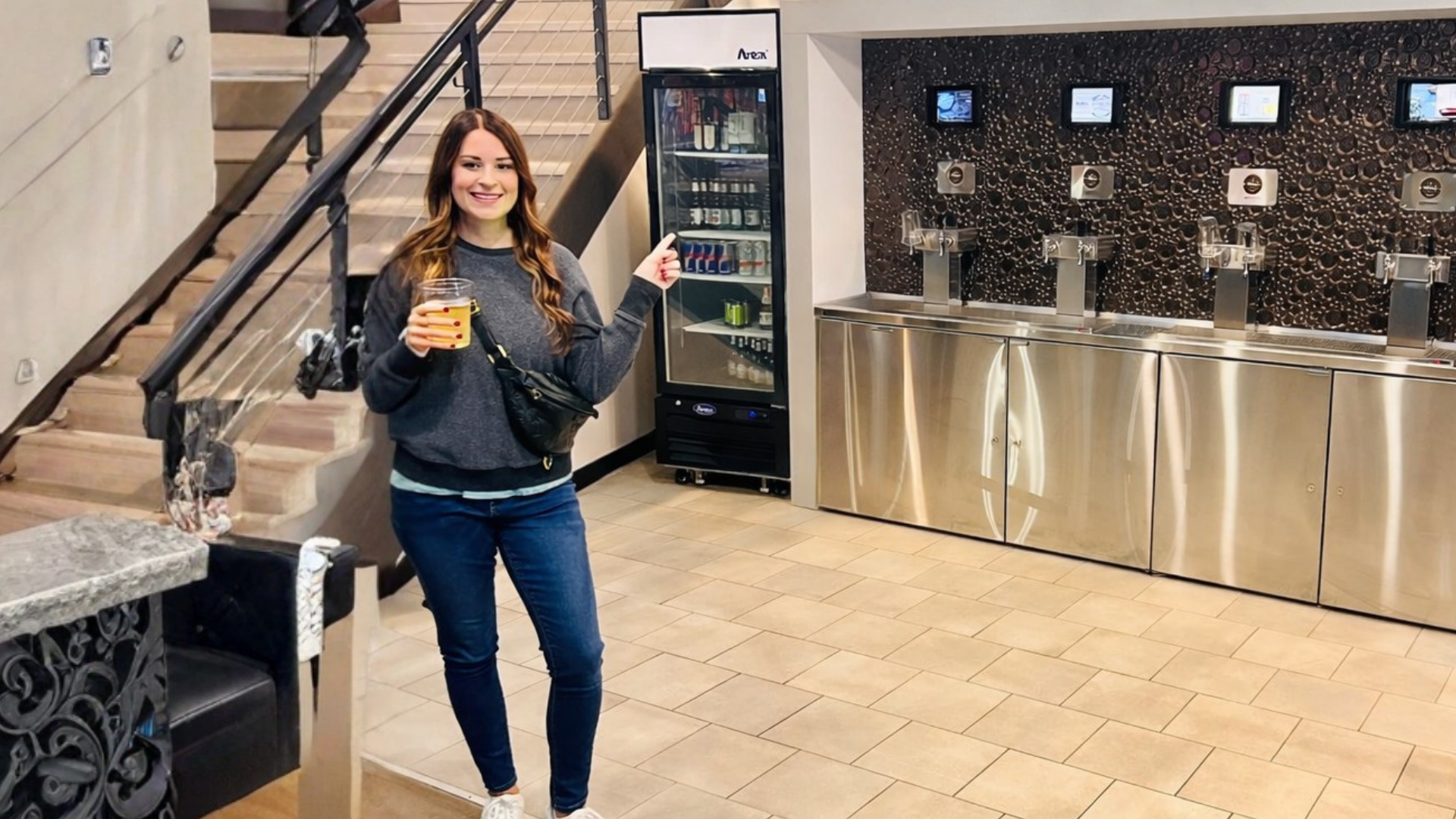 Woman in a gray sweater and jeans holding a drink, standing in front of a beverage machine in a commercial or office break room.