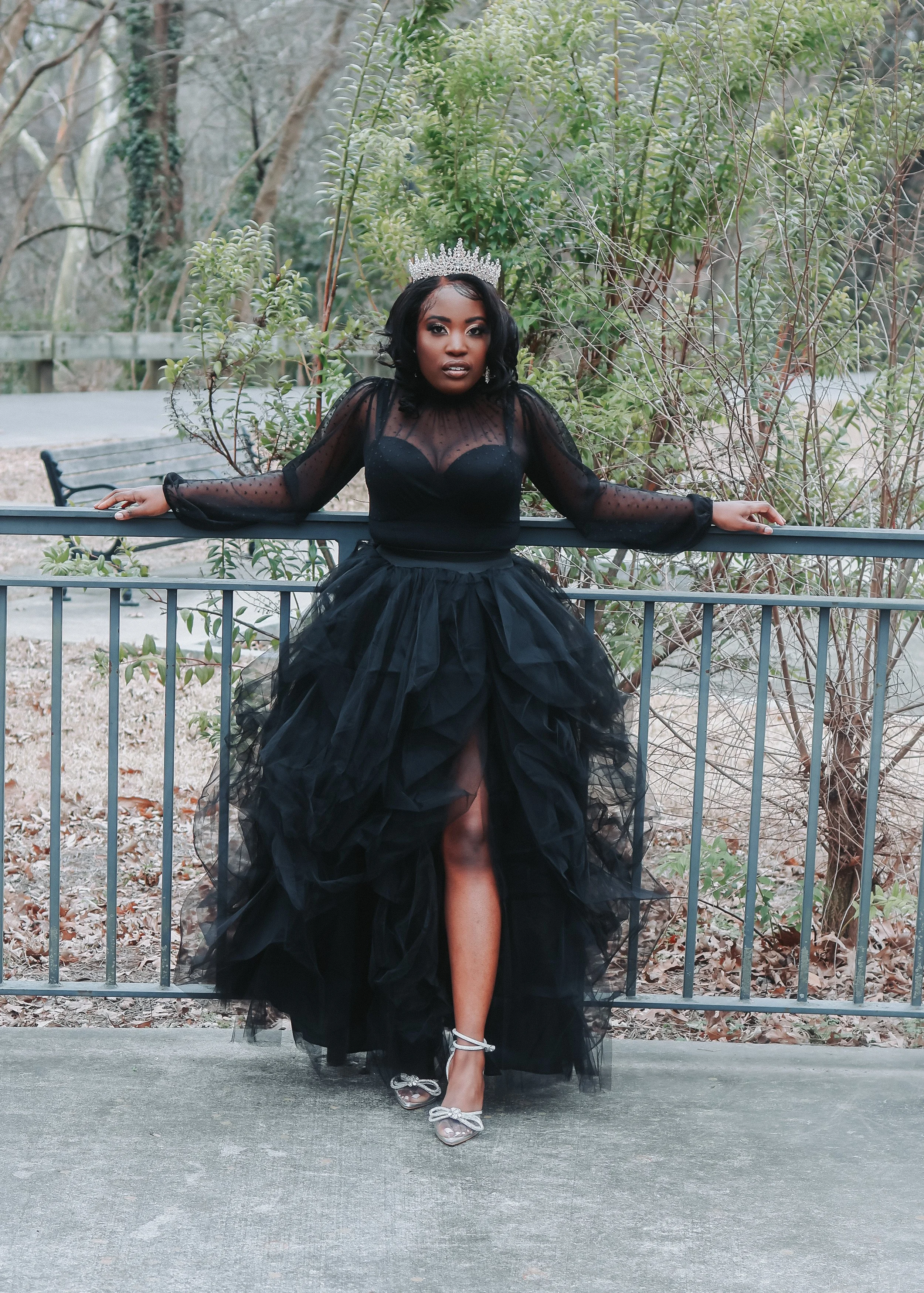 A woman wearing a black tulle gown and a tiara, standing outdoors behind a metal railing, with trees and bushes in the background.