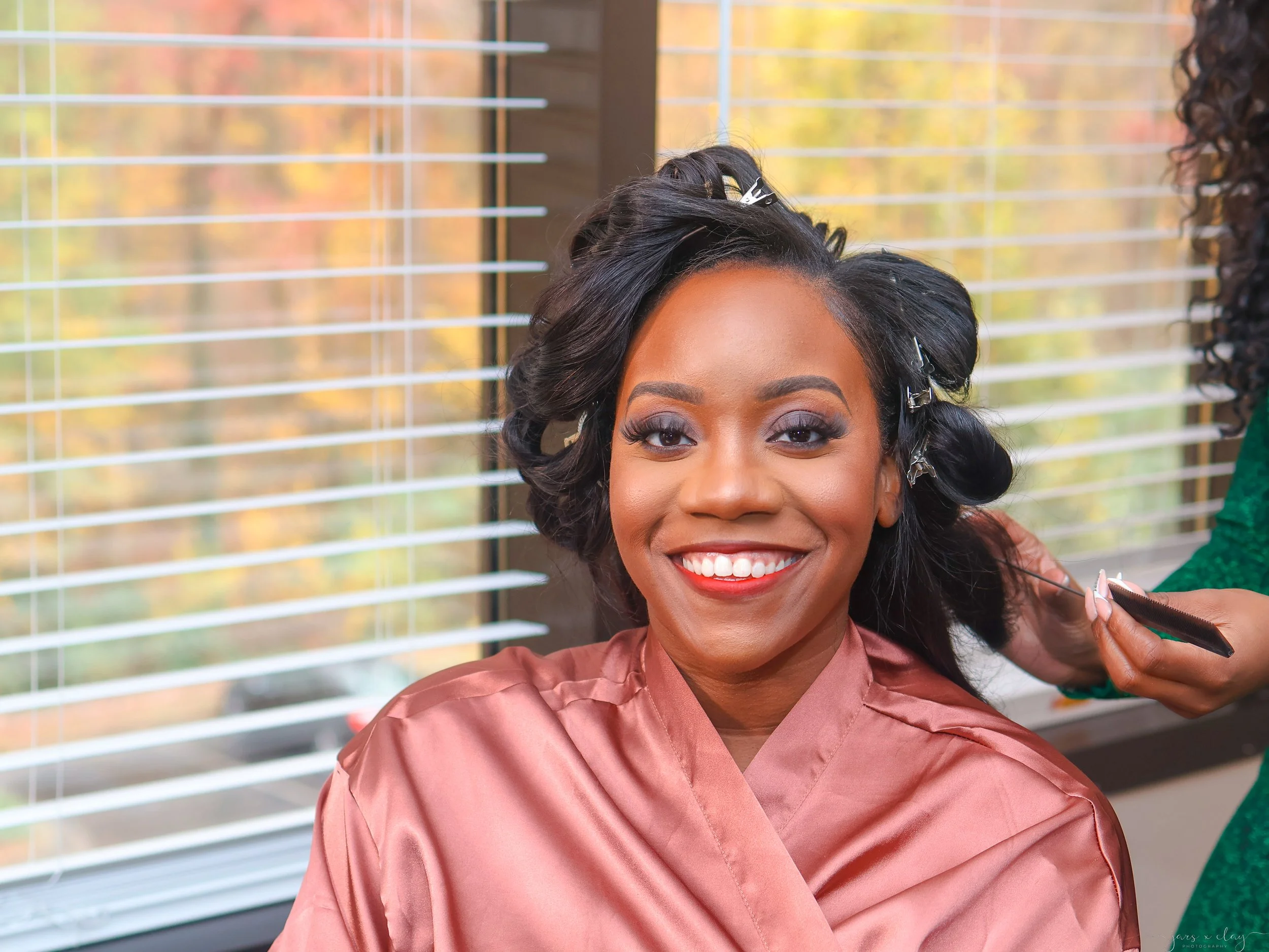 A woman smiling, getting her hair styled, wearing a peach-colored satin robe, with curlers in her hair, sitting in front of window blinds with fall outdoor scenery visible through the window.
