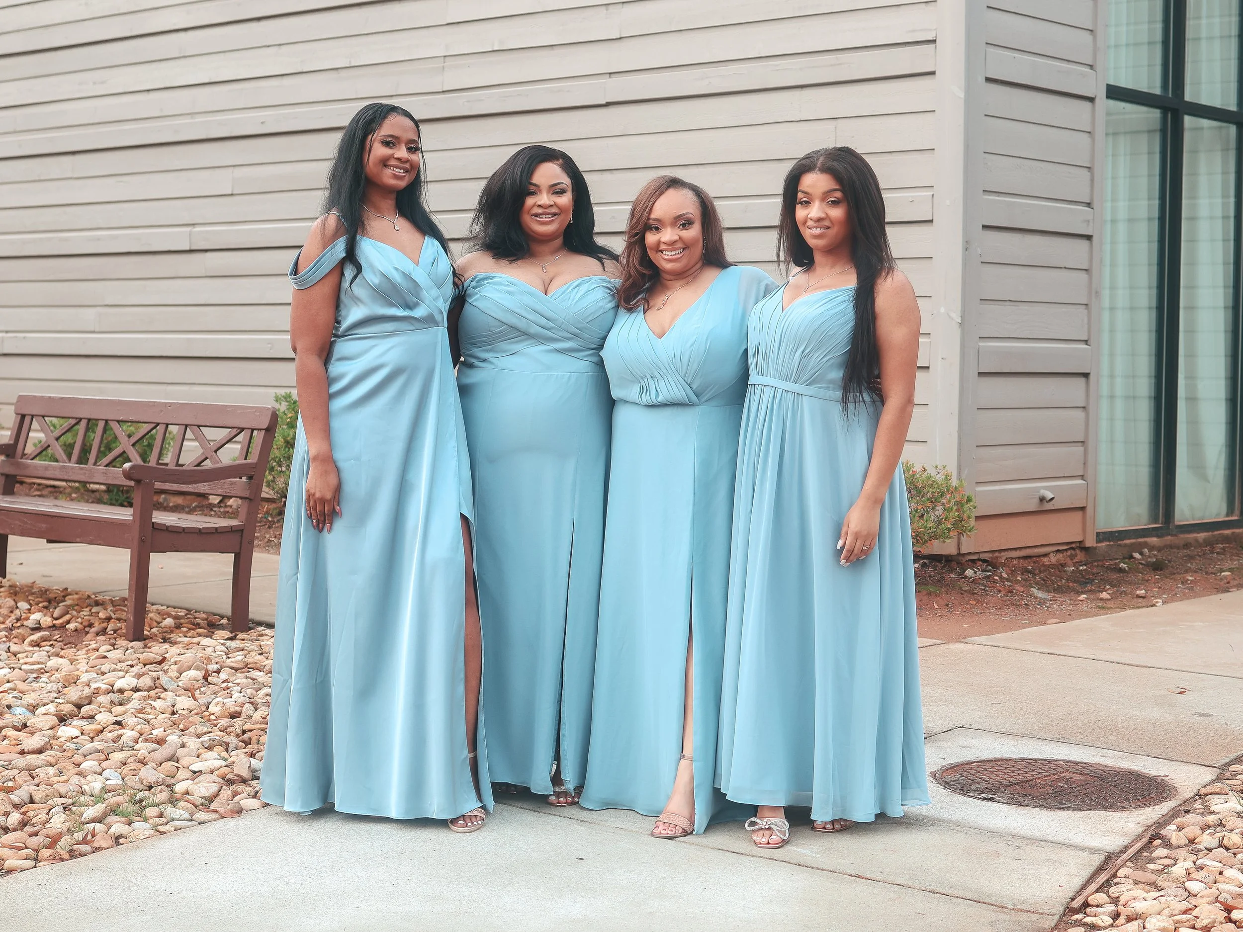 Four women in matching light blue evening gowns standing outside in front of a beige building with horizontal siding. They are smiling and posing for the photo.