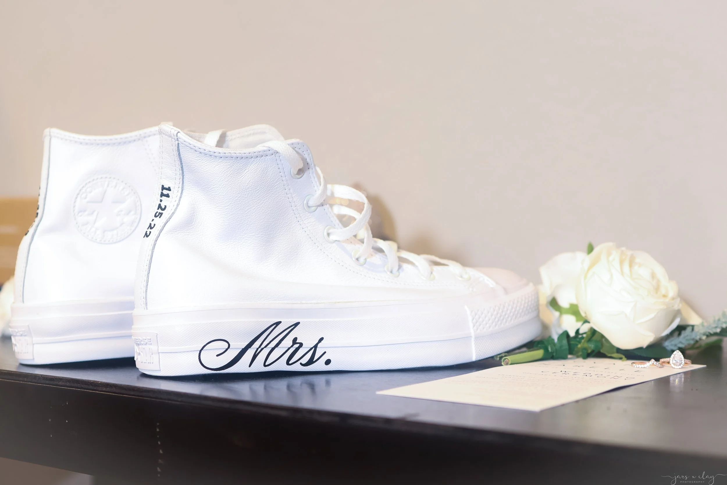 A white high-top Converse sneaker with the words 'Mrs.' written on the side of the sole, likely part of a bridal-themed display, with white flowers nearby.