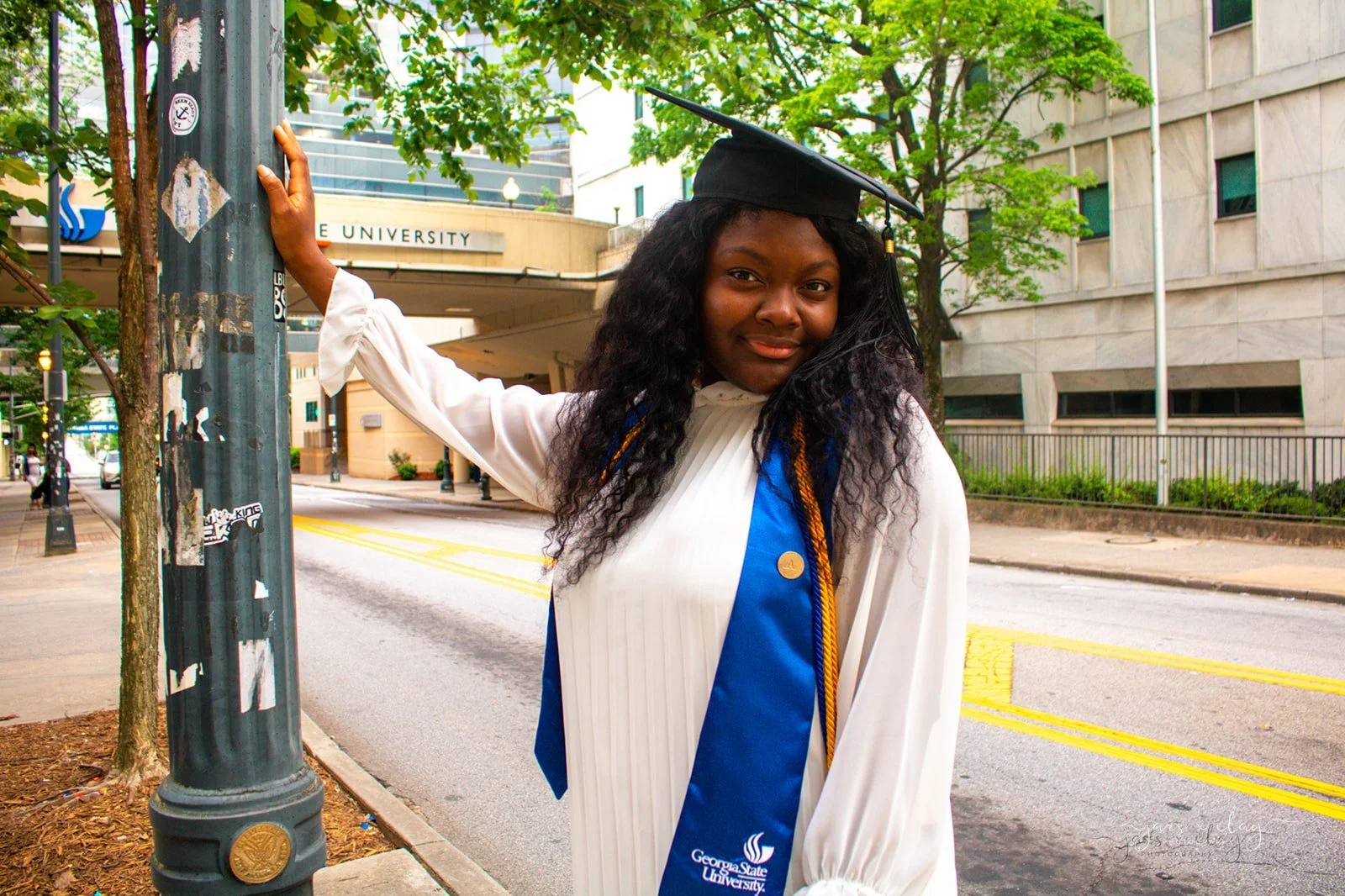 A young woman in a graduation gown and cap standing on a city sidewalk, smiling and holding onto a lamppost, with trees and a university building in the background.