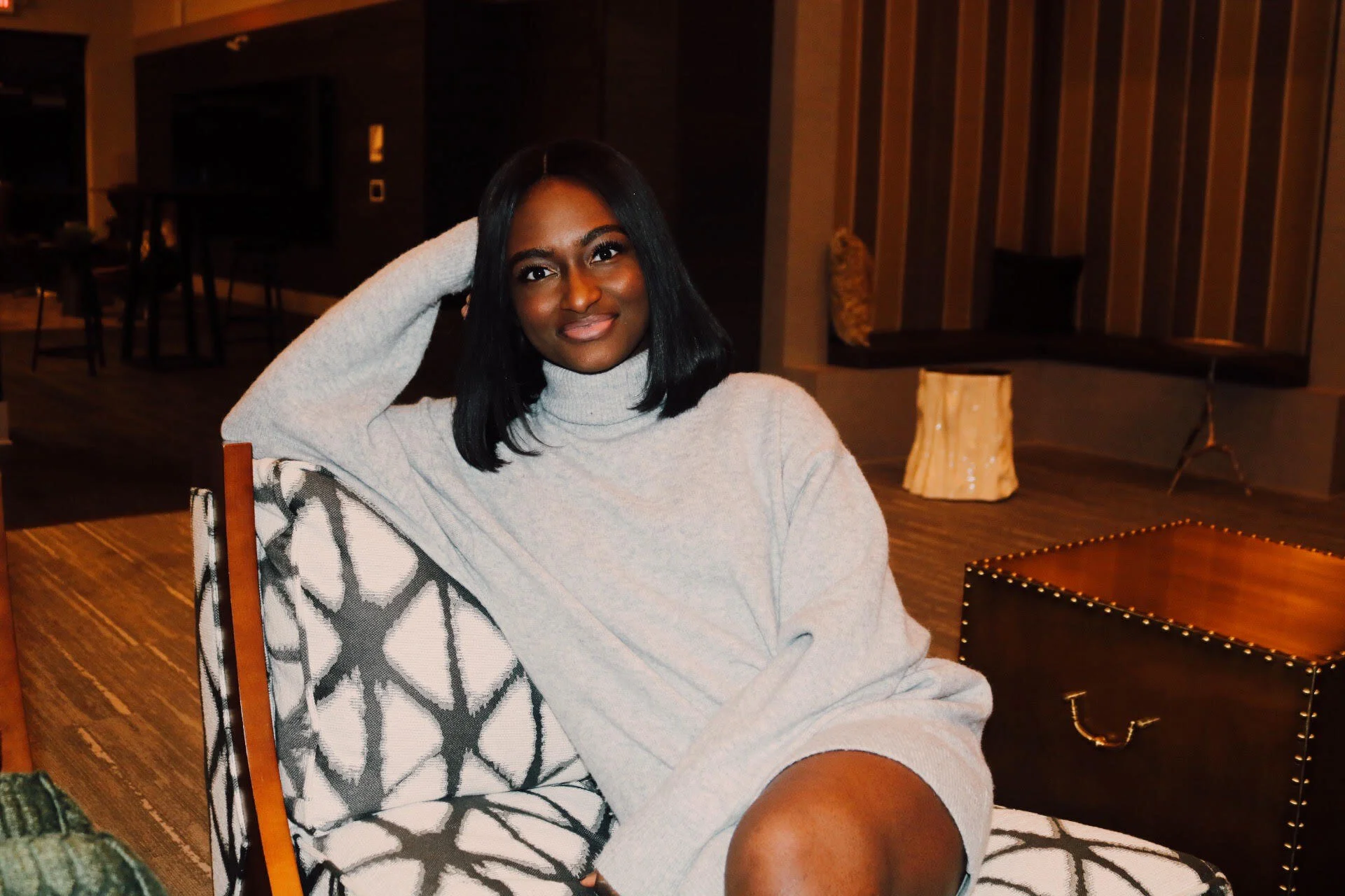 A woman with black hair wearing a light gray turtleneck sweater sitting in a cozy, modern indoor setting with wooden accents and a decorative end table.