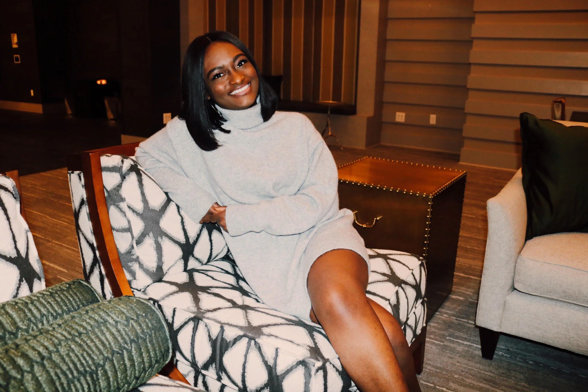 A smiling woman with medium skin tone and shoulder-length black hair, wearing a light gray turtleneck sweater dress, sitting on a black and white patterned couch in a modern living room with wood-paneled walls and warm lighting.