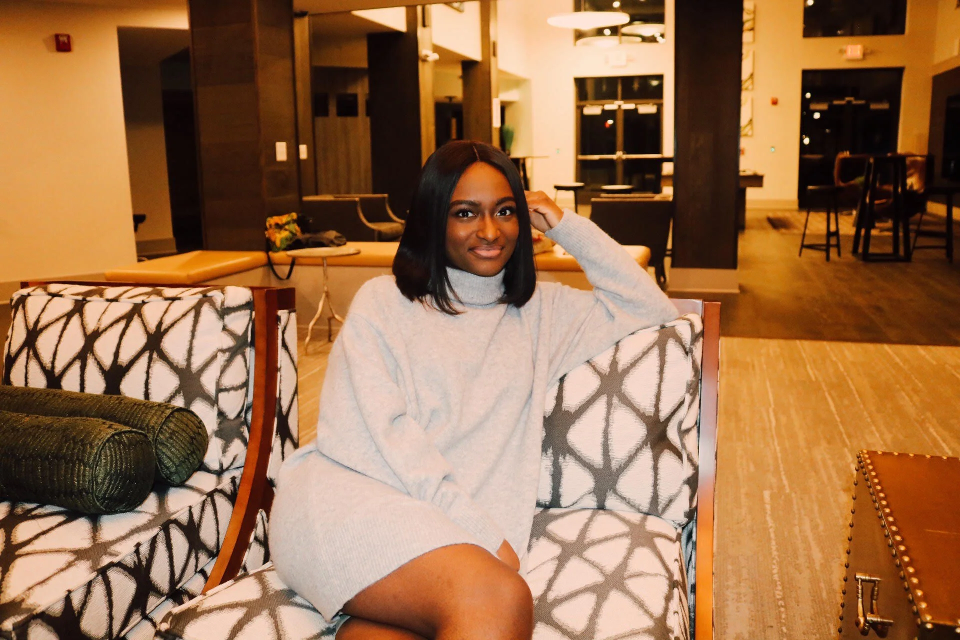 A woman with shoulder-length black hair sitting on a patterned chair in a warmly lit lobby area, wearing a light gray turtleneck sweater.