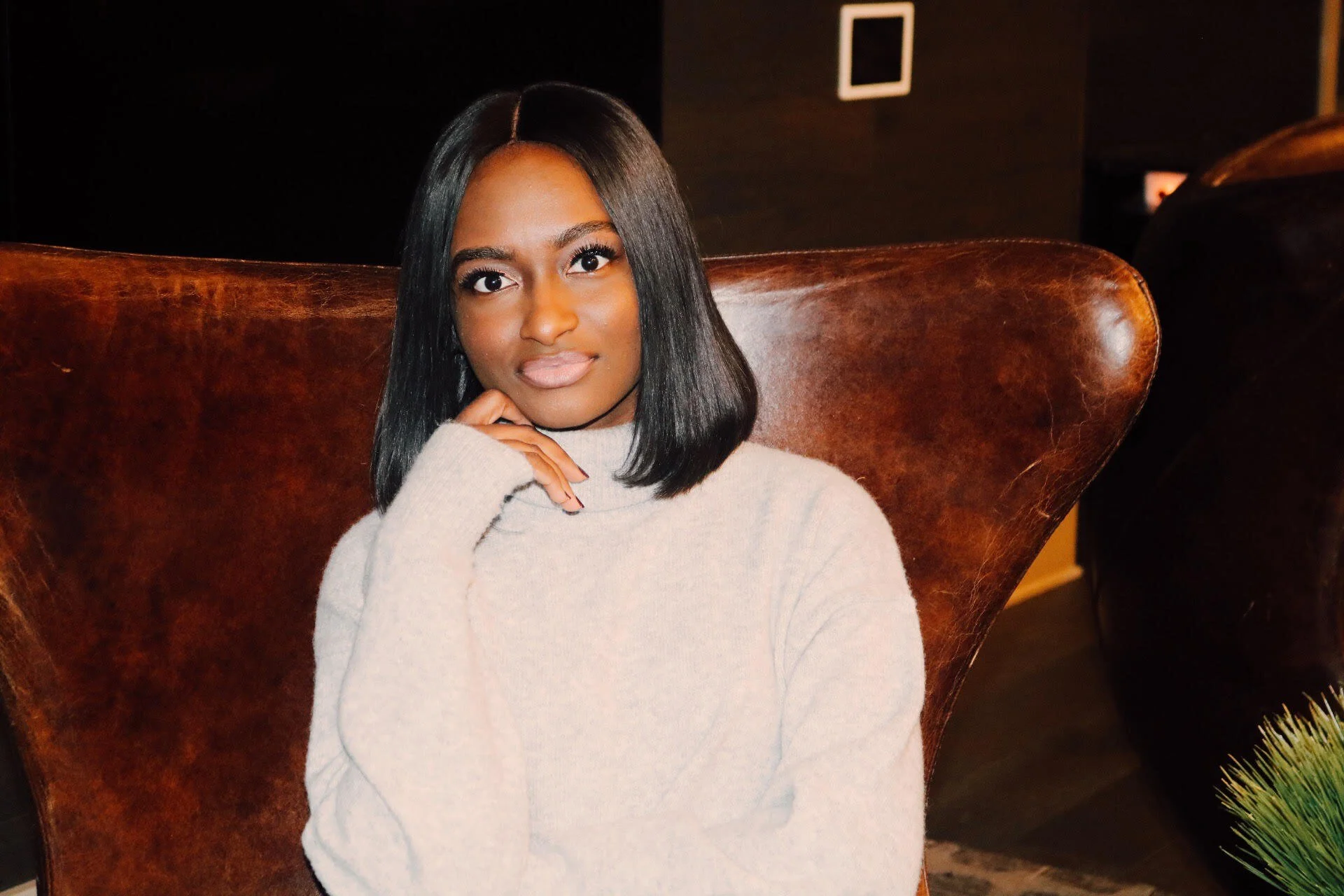 Young woman with straight black hair, sitting on a brown leather chair, wearing a light beige turtleneck sweater, in a dimly lit room.