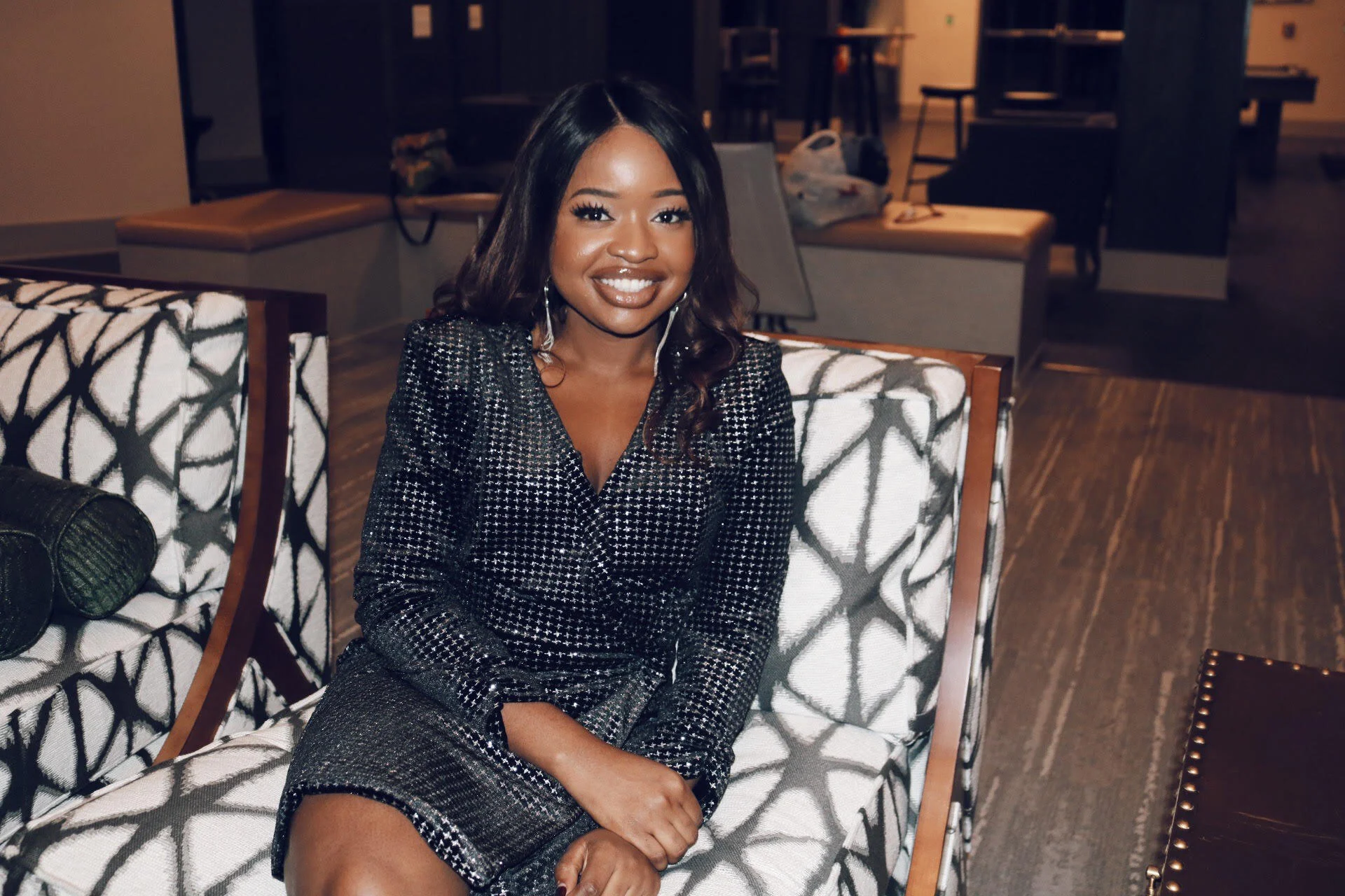 A young woman smiling and sitting on a patterned armchair in a dimly lit room, wearing a black and white dress with a checkered pattern.