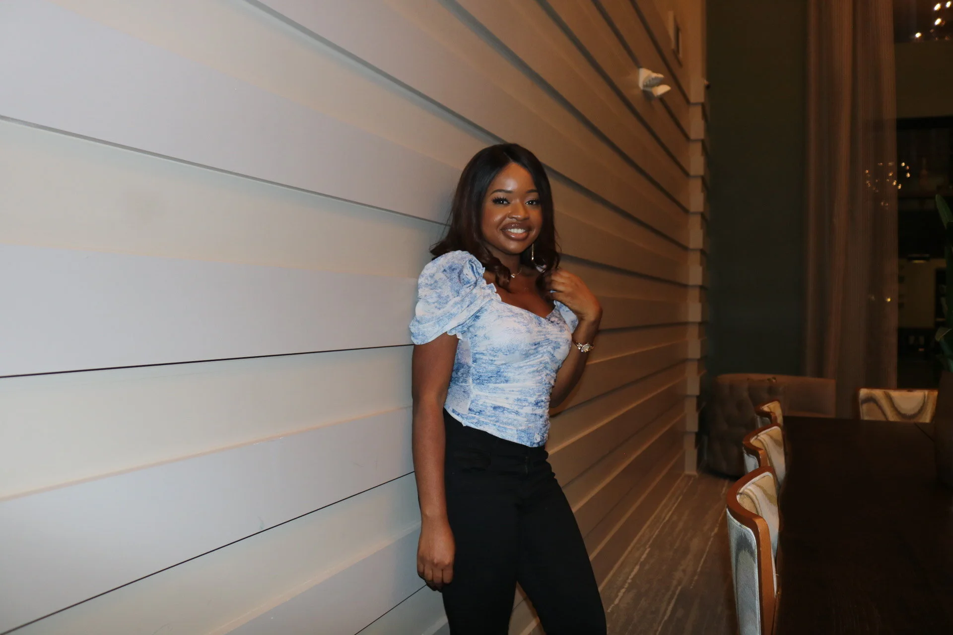 A woman with dark hair styled in loose waves smiling and standing next to a beige wood-paneled wall in a warmly lit interior space, wearing a blue patterned short-sleeve top and black pants.