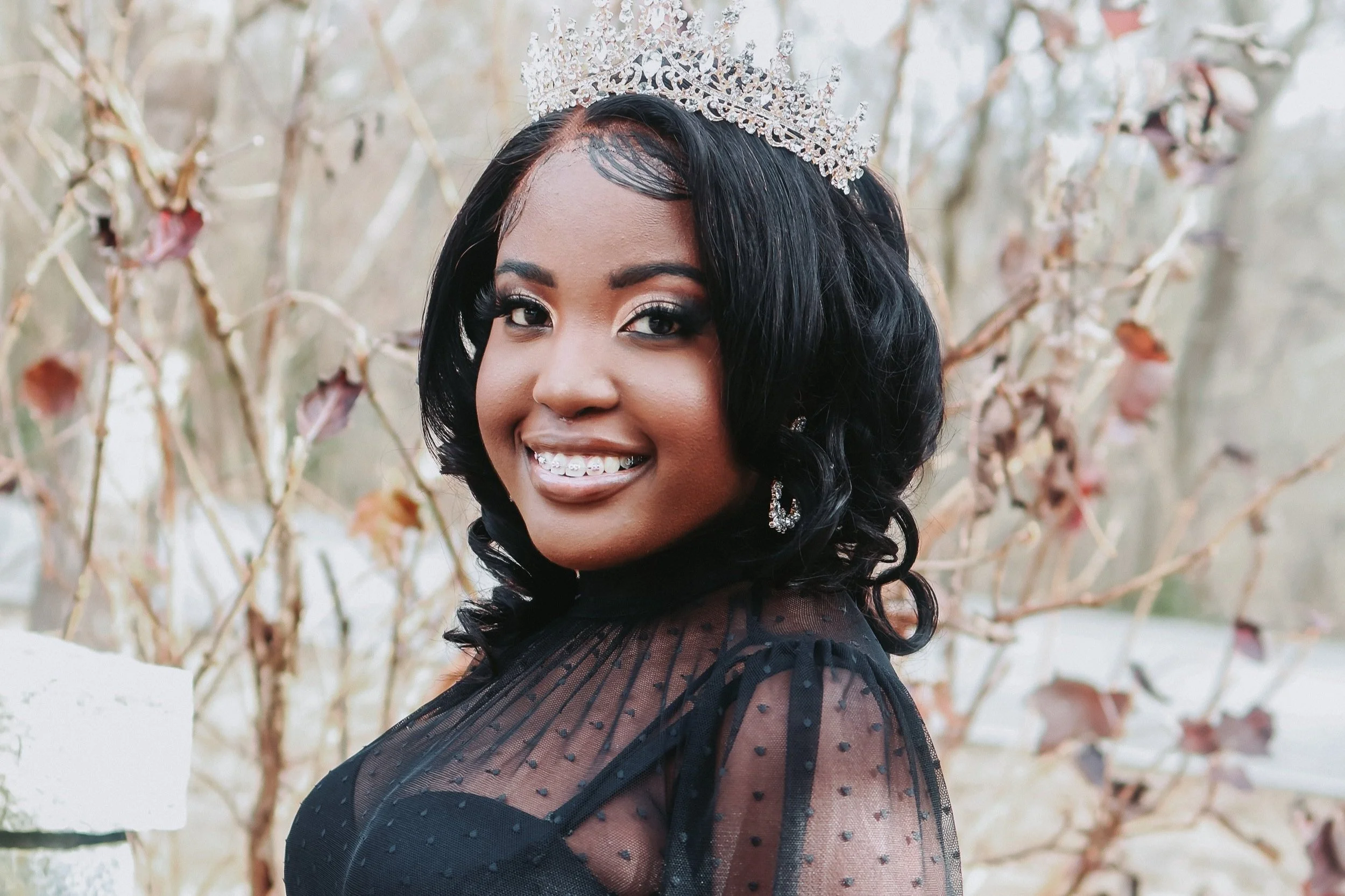 A woman with dark hair, wearing a sparkling crown, smiling with braces, and dressed in a black, sheer polka-dot top, standing outdoors in front of a tree with dried leaves.